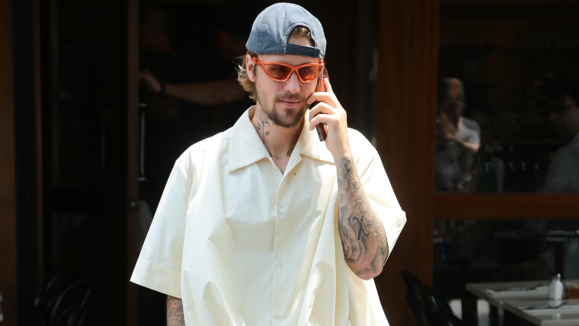 Justin Bieber with tattoos, wearing a light shirt, orange sunglasses, and a backward cap, talks on the phone while walking outside.