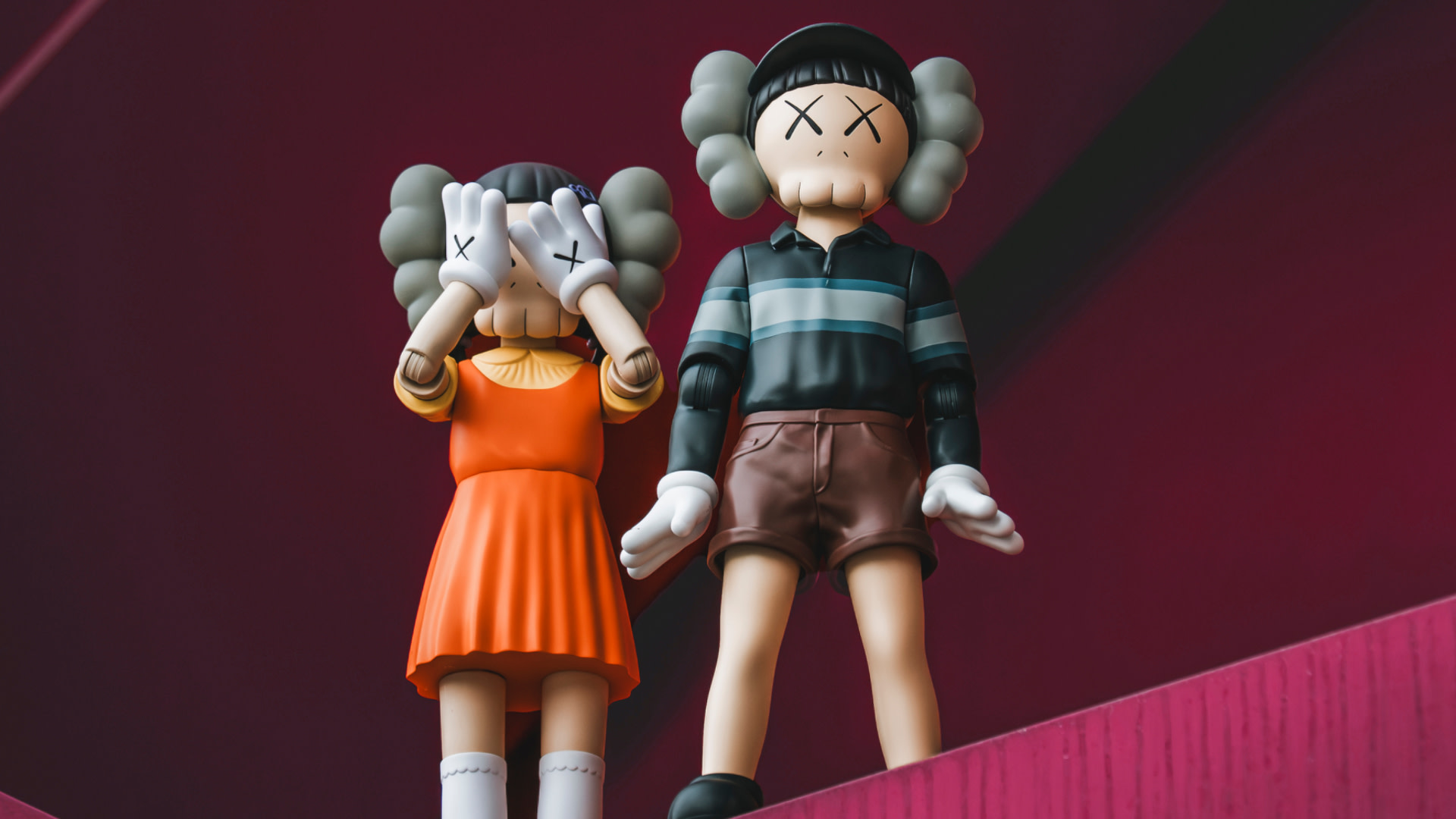 Two KAWS figures with X eyes, one in an orange dress covering its eyes, the other in a striped shirt and shorts, against a red background.