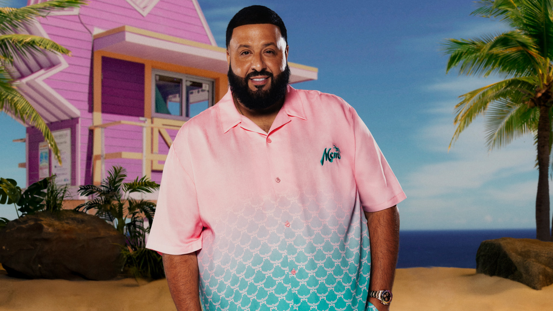 DJ Khaled stands on a beach, wearing a pink and blue shirt, with a pink house and palm trees in the background.