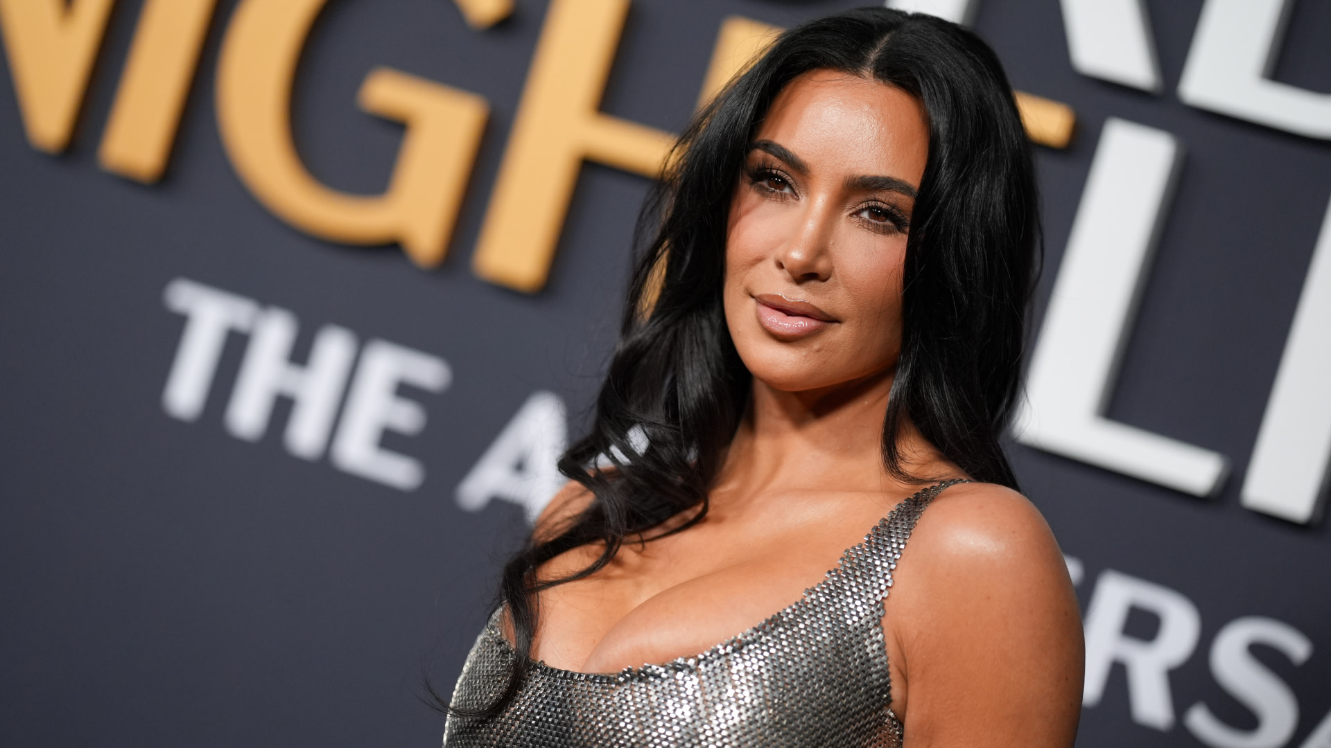 Kim Kardashian with long, dark hair in a silver dress poses confidently on a red carpet with a dark backdrop featuring yellow and white text.