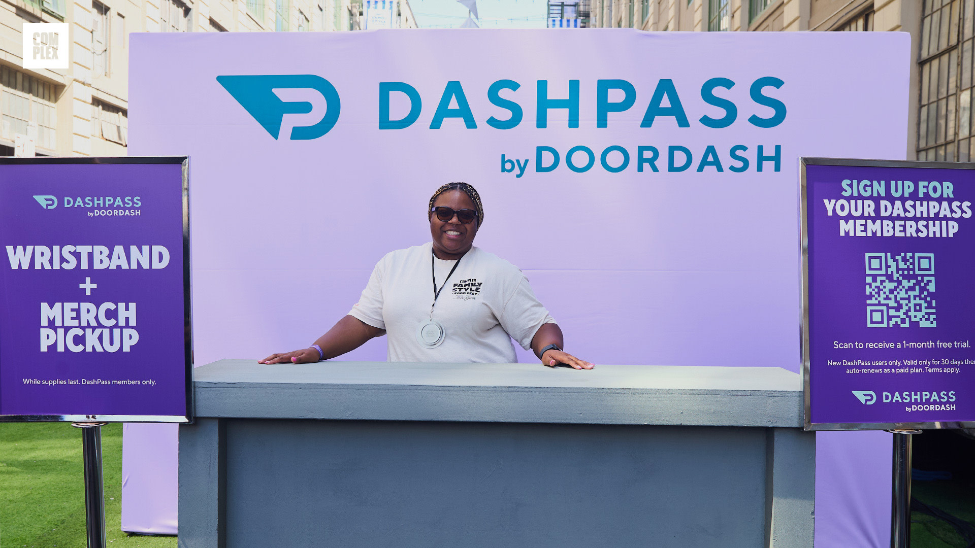 A person stands smiling at a DashPass by DoorDash booth, wearing sunglasses and a white shirt. Signs for wristband pickup and membership are visible.