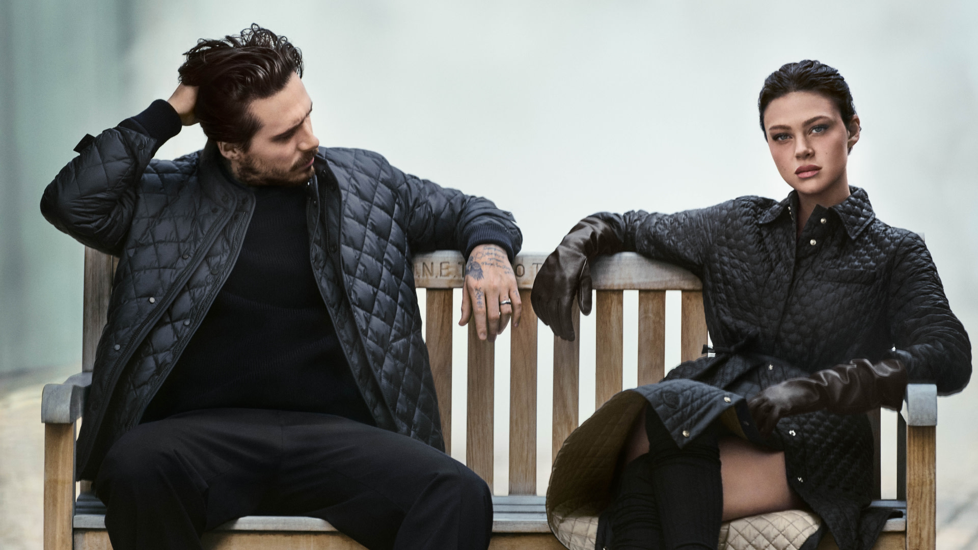 A man and woman in stylish quilted jackets sit on a bench, with the man touching his hair and the woman looking directly ahead.