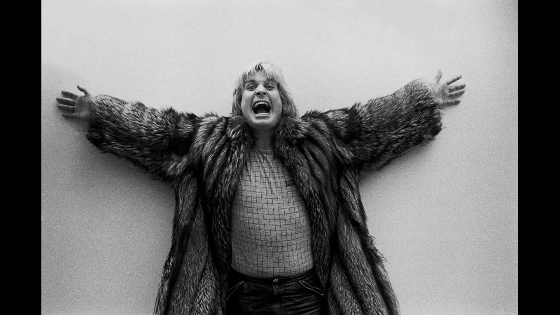 Ozzy Osbourne stands with arms outstretched, wearing a fur coat and patterned shirt, appears to be shouting in an expressive pose.