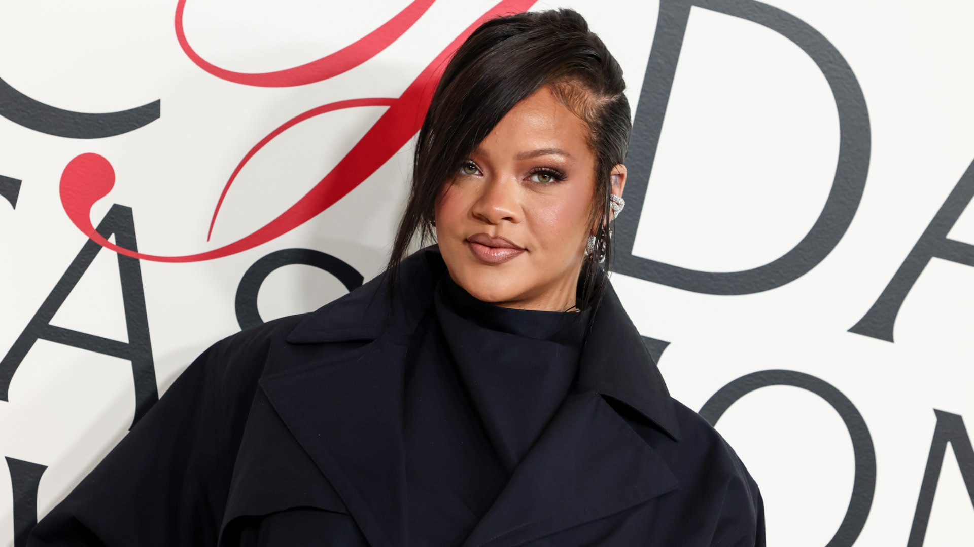 Rihanna at a fashion event, wearing a dark outfit with a side-swept hairstyle, standing in front of a stylized background.