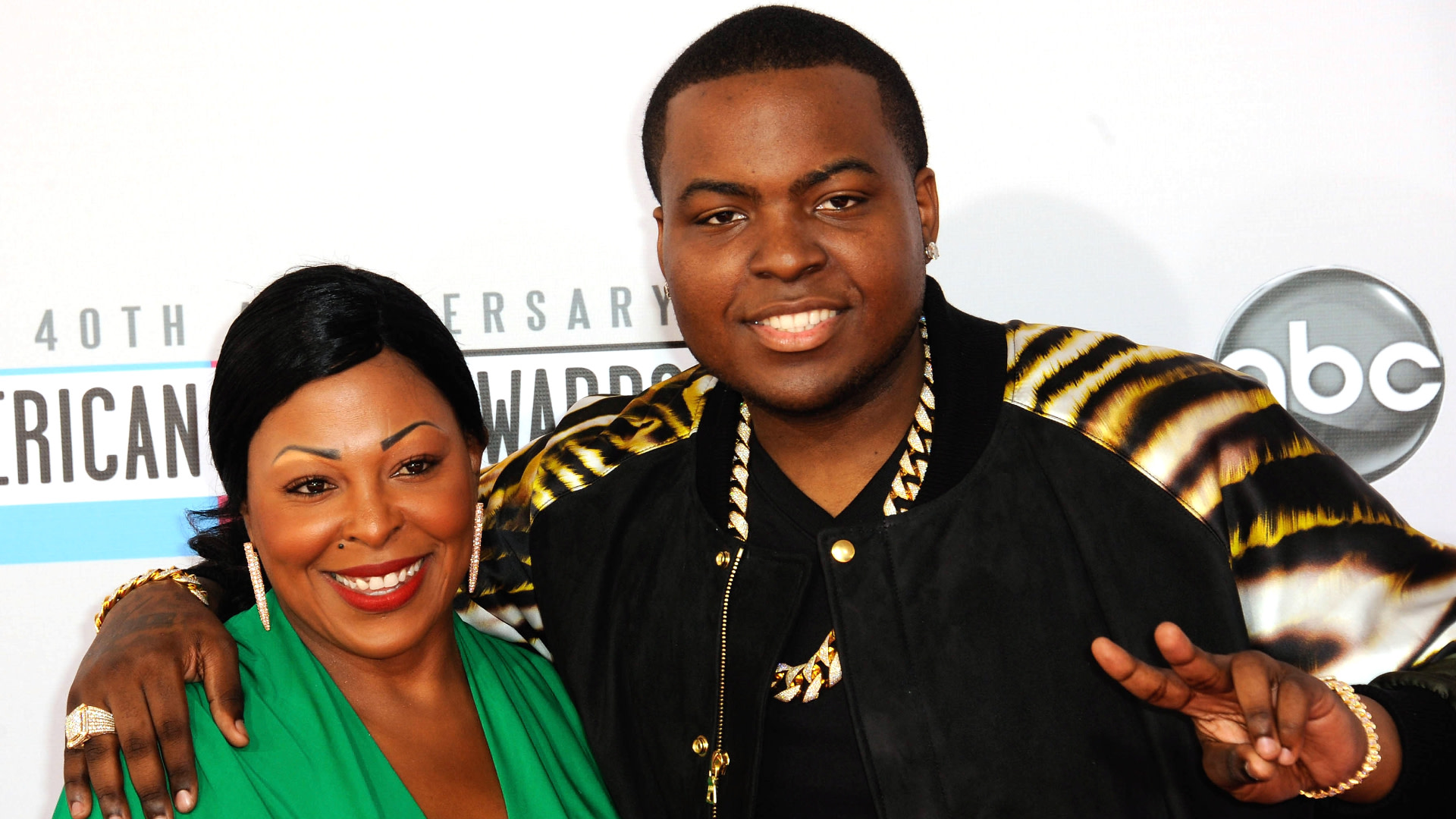 Sean Kingston and his mother smiling on the red carpet at the 40th American Music Awards. The man is wearing a black and gold jacket.