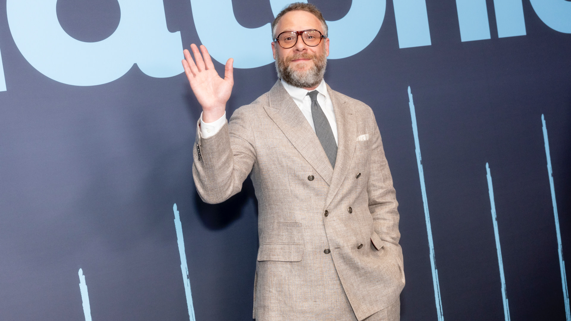 Seth Rogen in a light gray suit, white shirt, and tie waves at an event, standing against a dark blue backdrop with large text.