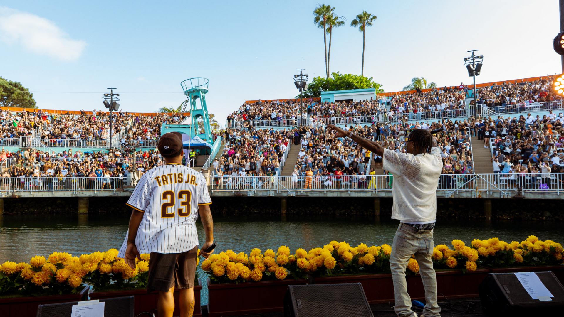 Bow Wow and Soulja Boy on stage facing a large audience near water. One wears a "Tatis Jr. 23" jersey. Bright yellow flowers line the stage.