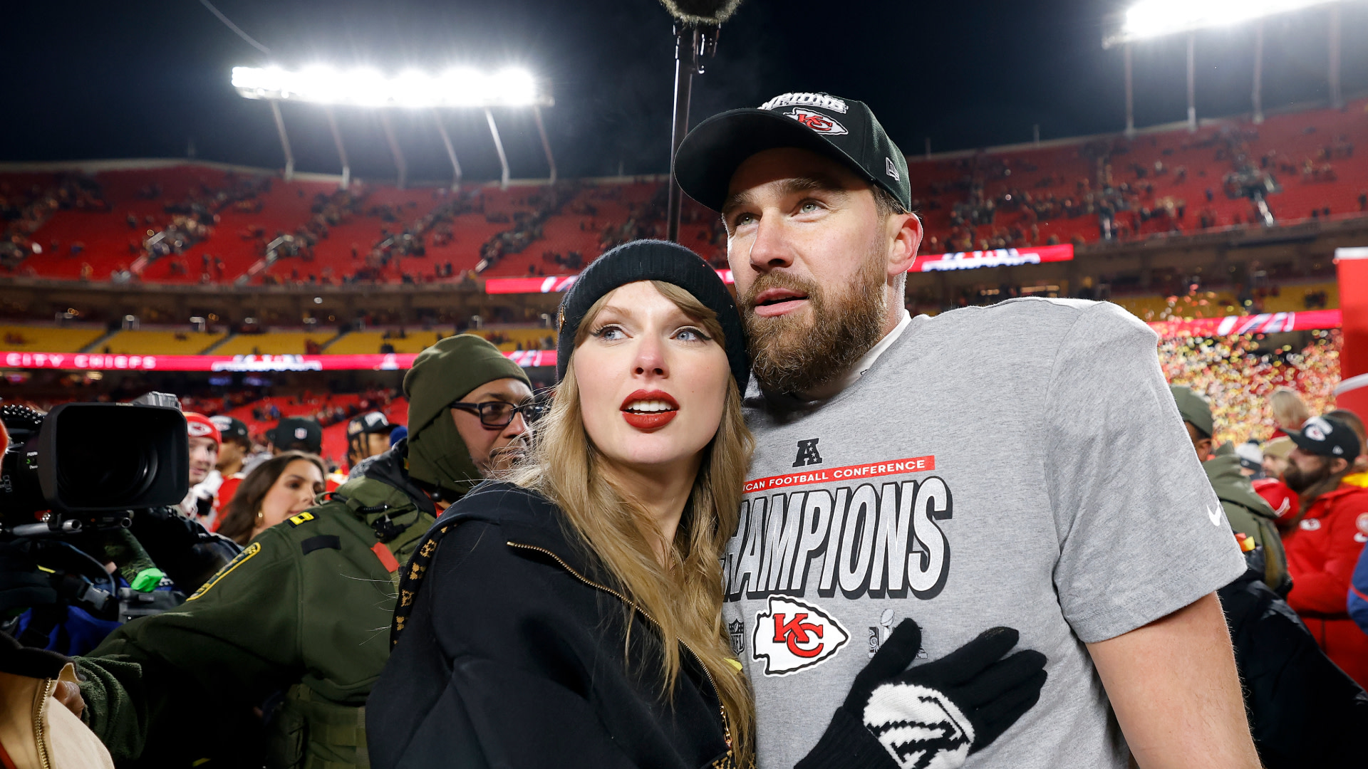 Taylor Swift and Travis Kelce at a Kansas City Chiefs game, celebrating on the field. Swift is wearing a black beanie and Kelce a cap.