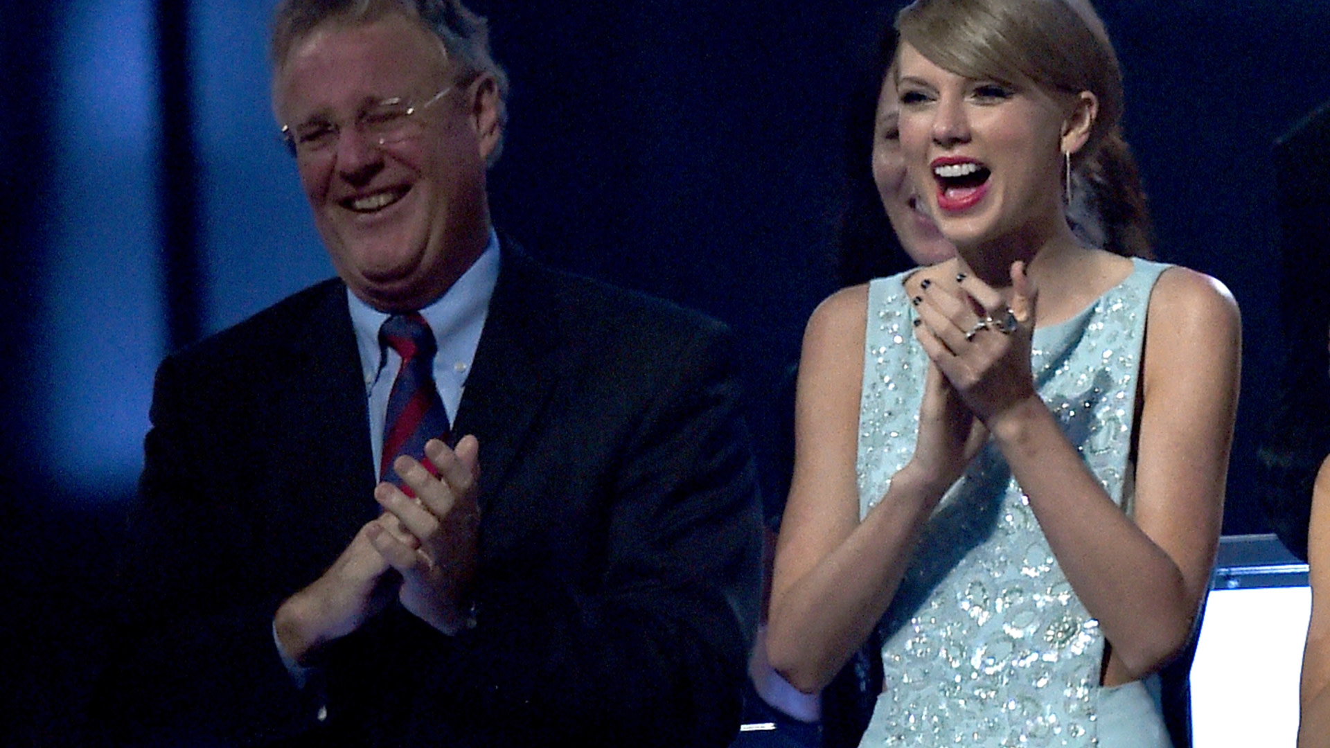 Scott Swift and Taylor Swift, wearing a light blue dress, are clapping and smiling at an event.
