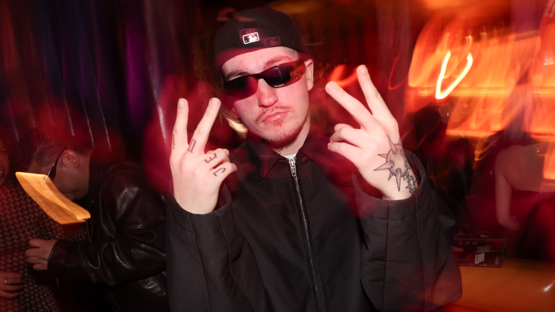 Tommy Richman in a dark jacket, cap, and sunglasses poses with peace signs in a dimly lit room with red and orange lights.