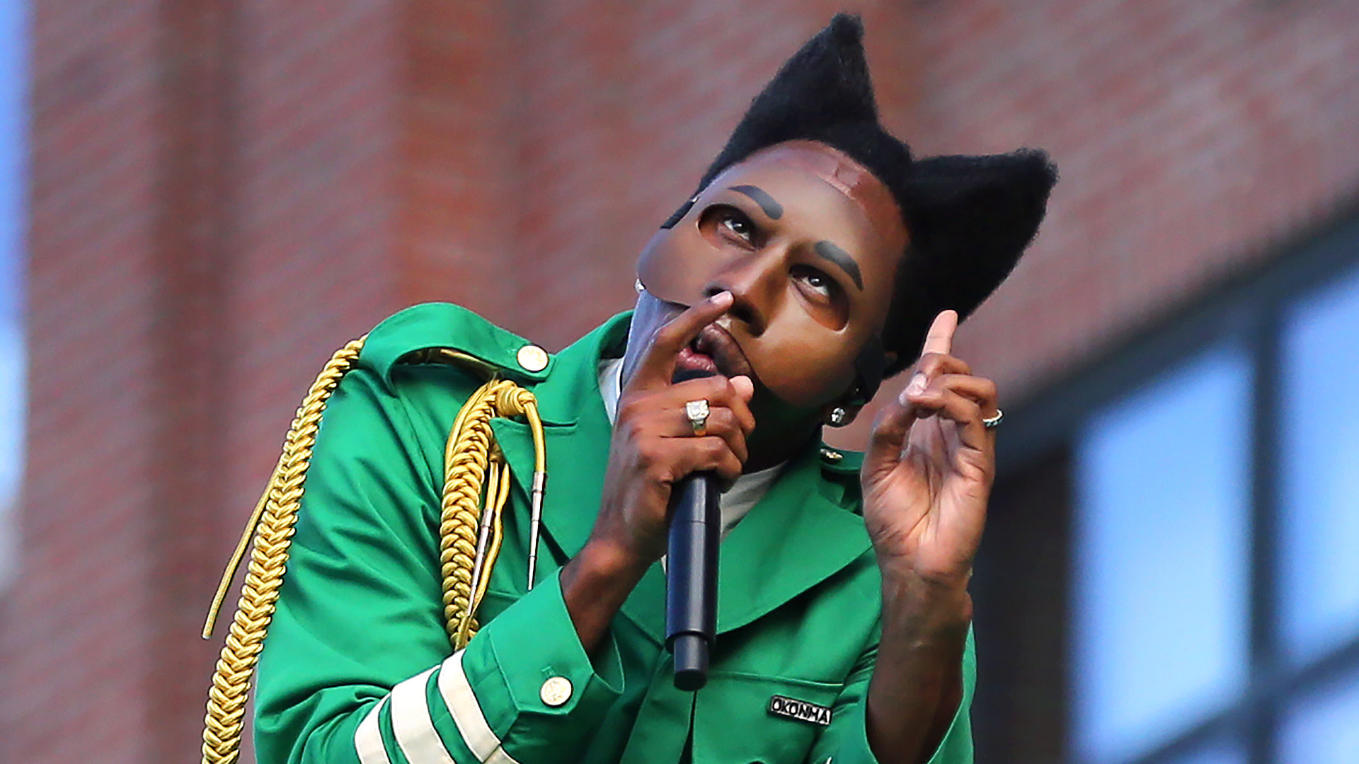 Tyler, The Creator in a green jacket with gold accents, wearing a unique mask and cat-ear hairstyle, holds a microphone on stage.
