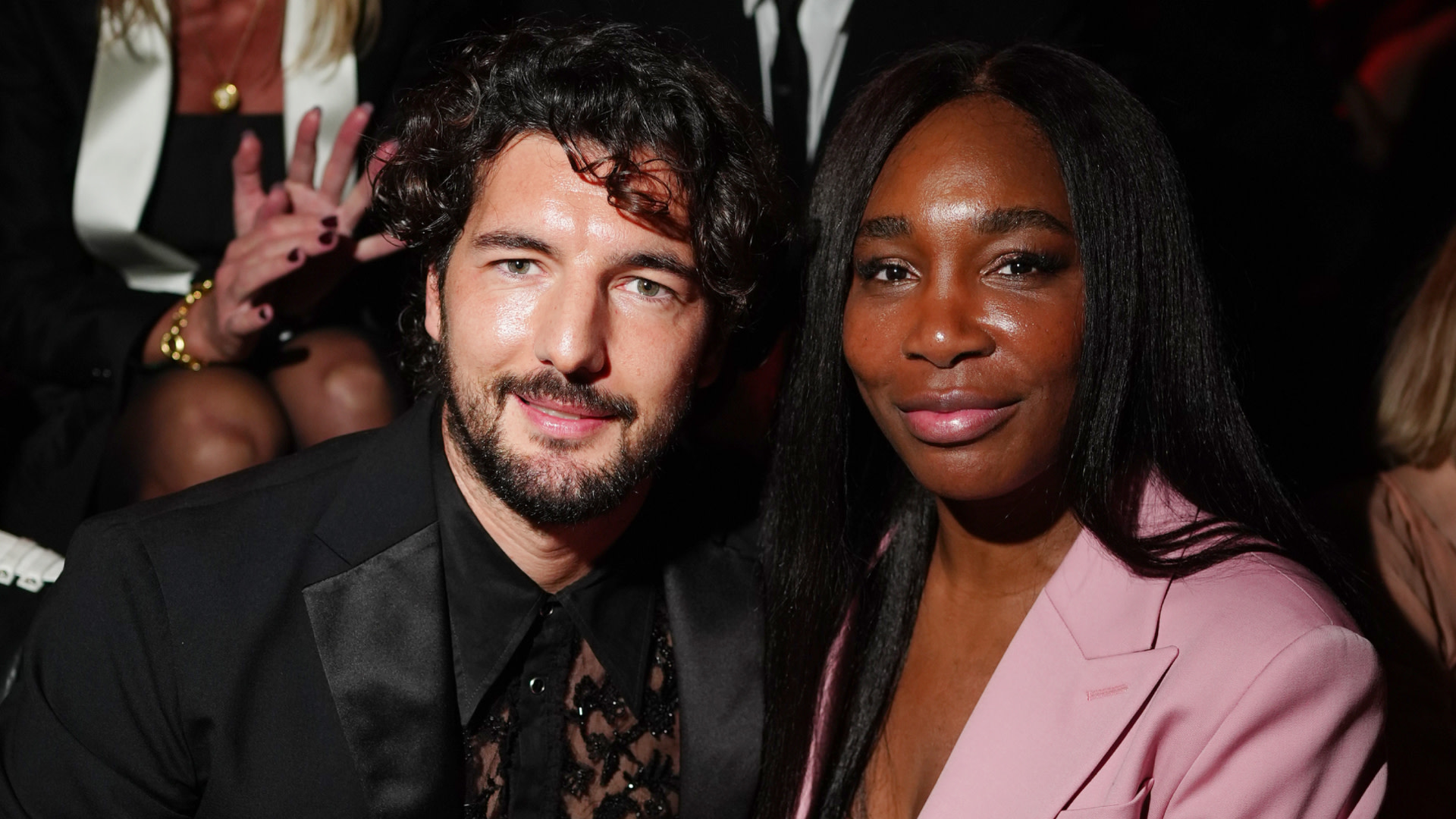 Andrea Preti in a black suit sits beside Venus Williams, who is wearing a pink blazer. They are at an indoor event.