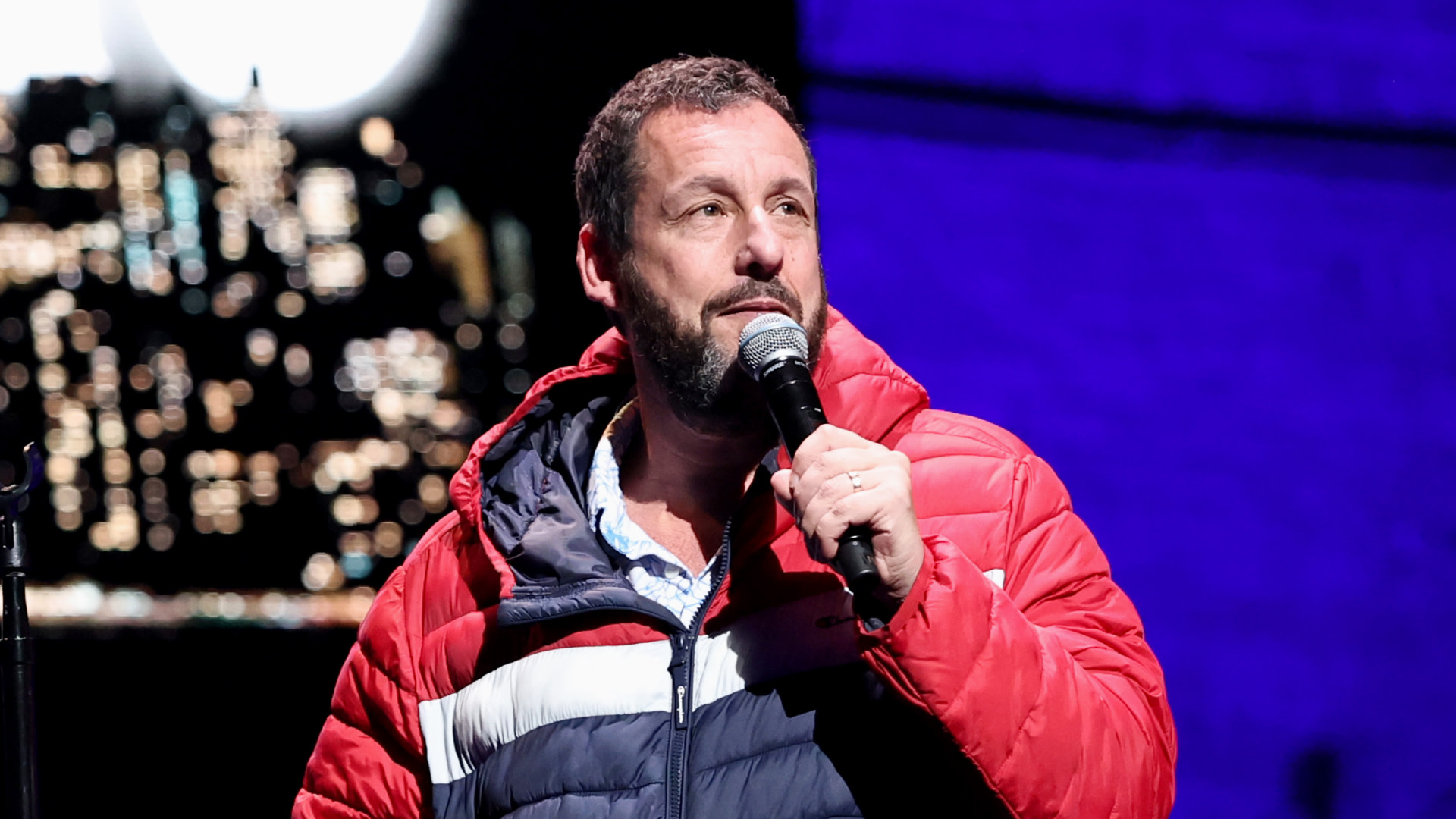 Adam Sandler performing on stage, wearing a red and blue jacket, holding a microphone.