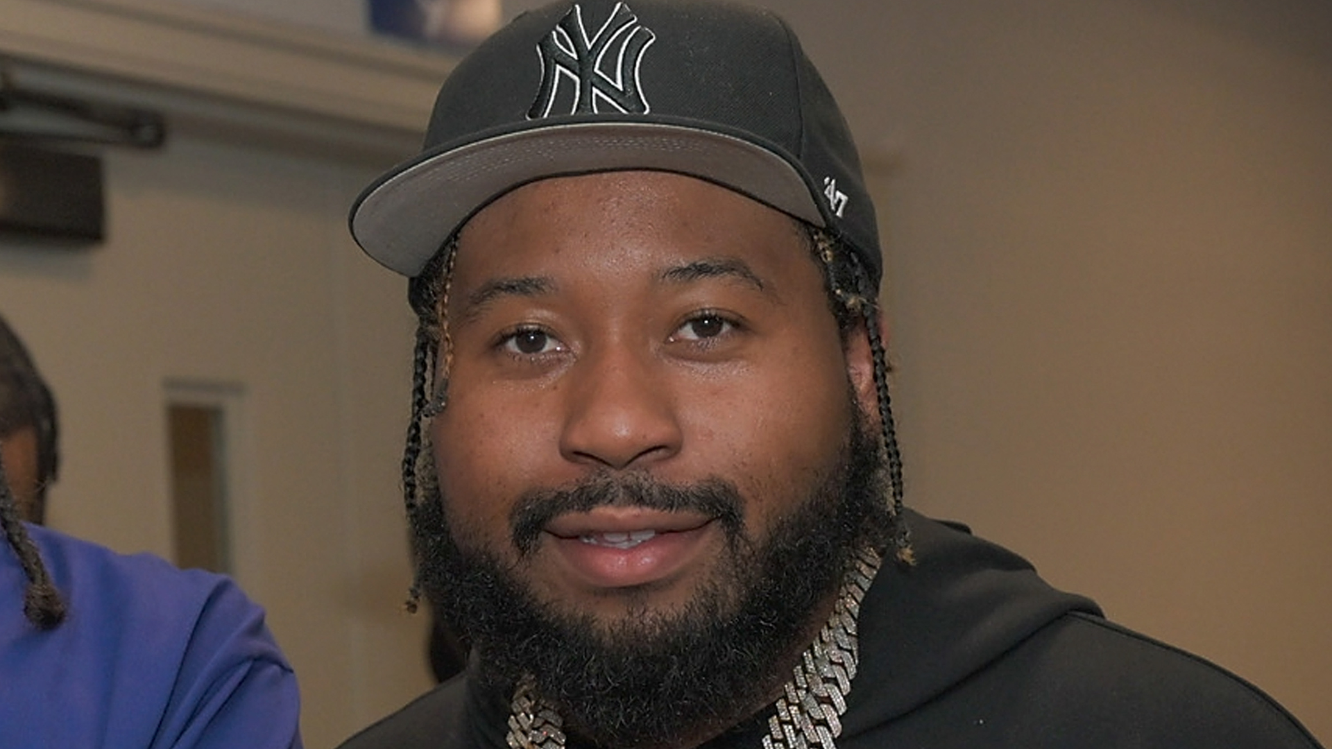 A man with a beard wearing a New York Yankees cap and a chain necklace, smiling indoors.