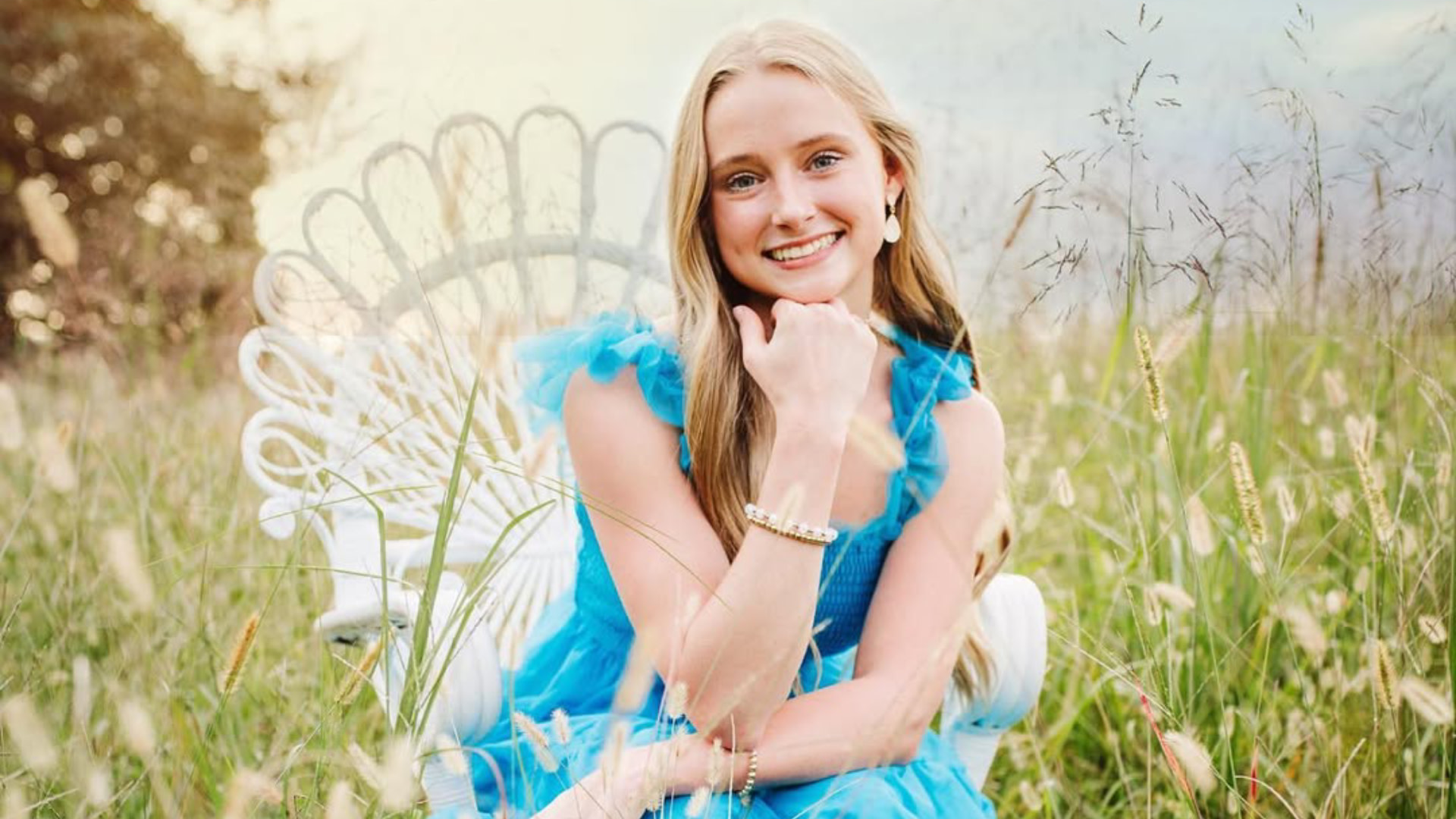 TikTok influencer Anna Grace Phelan in a blue dress sits on a white chair in a grassy field, smiling with her chin resting on her hand.