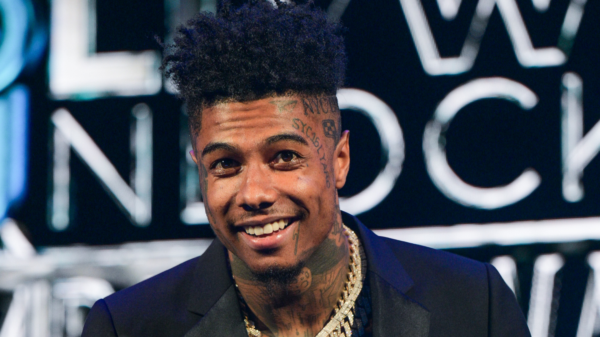A man with tattoos on his face and neck, wearing a dark blazer and gold chains, smiles at an event with a blurred background.