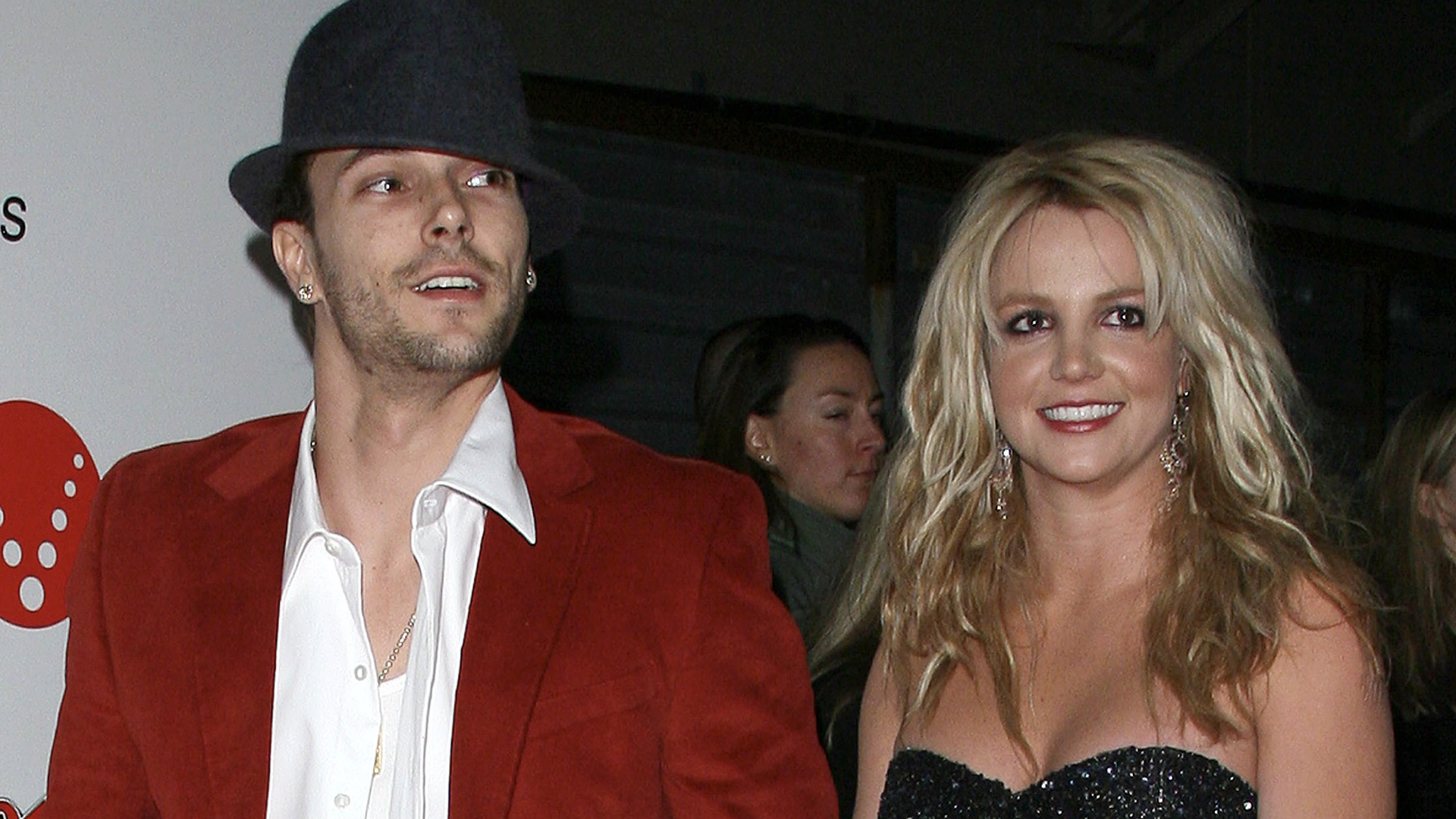 Kevin Federline in a red jacket and hat with Britney Spears in a black dress, smiling at an event.