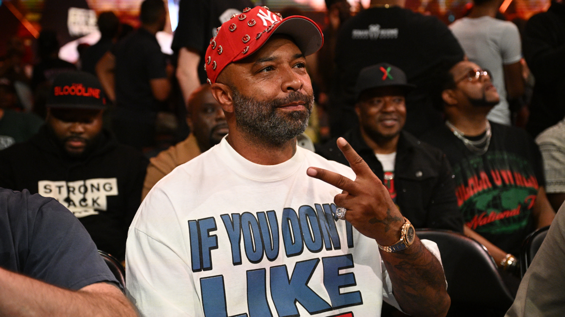 Joe Budden wearing a red cap and a white shirt with text, making a peace sign at an event. Others are seated around him.