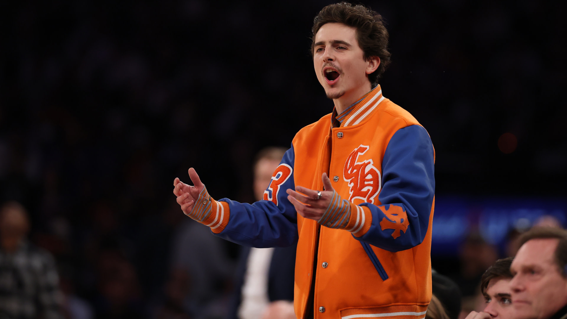 Timothee Chalamet in an orange and blue jacket is animatedly gesturing, possibly at a sports event.