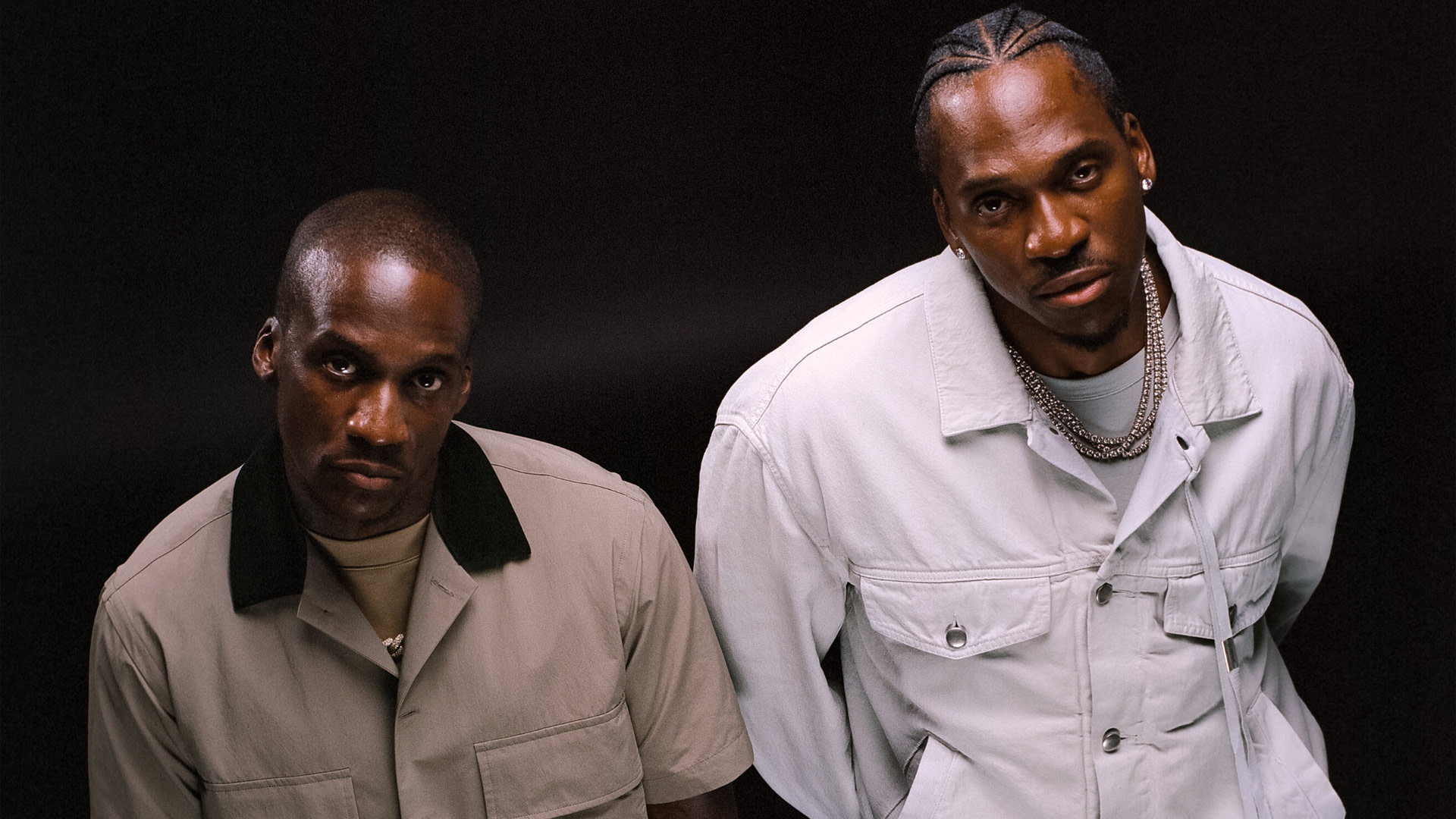 Clipse in stylish outfits pose against a dark background. Malice wears a beige jacket, Pusha T a white jacket with chains.
