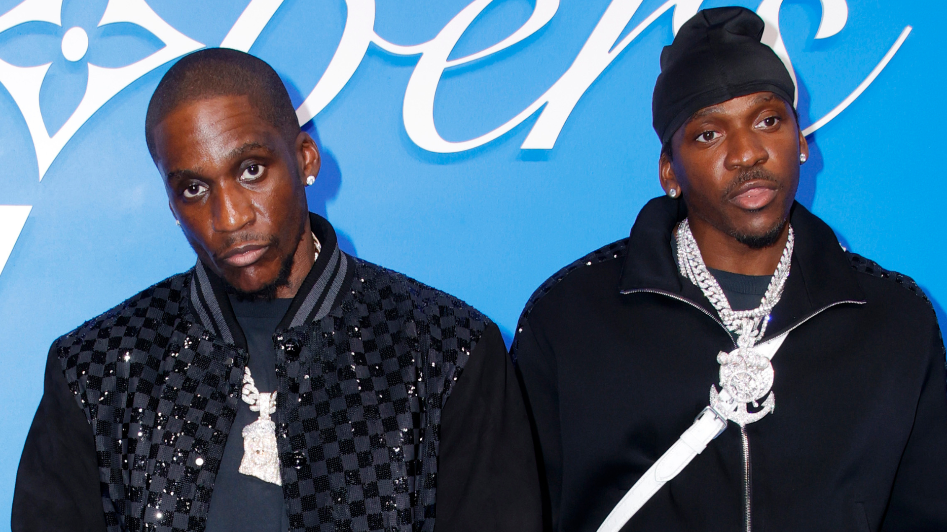 Two men in black, wearing diamond necklaces, stand against a blue background with white text.