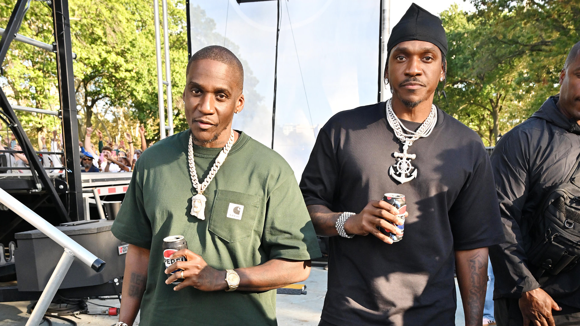 Malice and Pusha T standing outdoors, both holding drinks. One wears a green shirt, the other a black shirt and cap, both with chains.