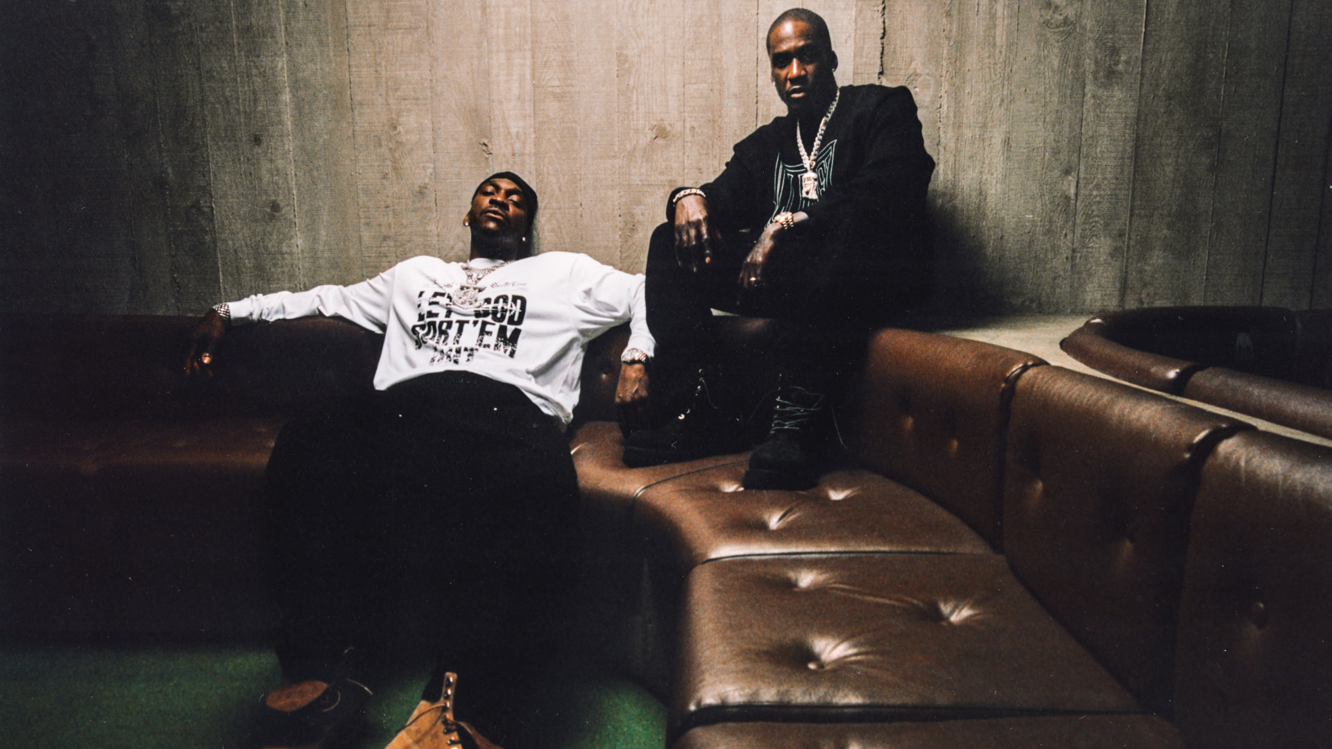 Clipse are sitting on a brown leather couch in a dimly lit room. Pusha T is reclining in a white shirt, Malice is sitting upright in black.
