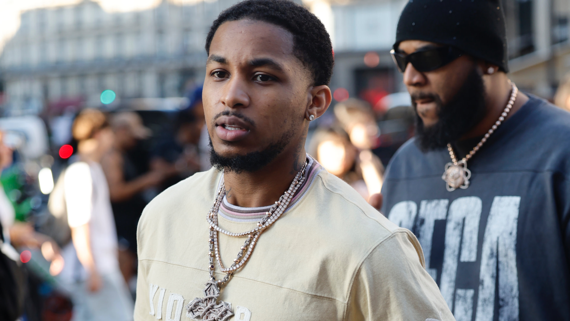 DDG wearing a beige shirt and multiple necklaces walks outside, with another man in sunglasses and a beanie in the background.