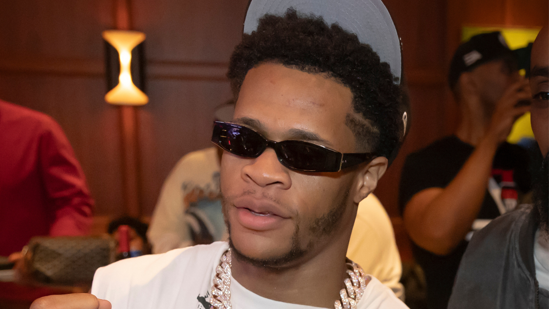 A person wearing sunglasses, a cap, and a chain necklace, with a blurred background of people and warm lighting.