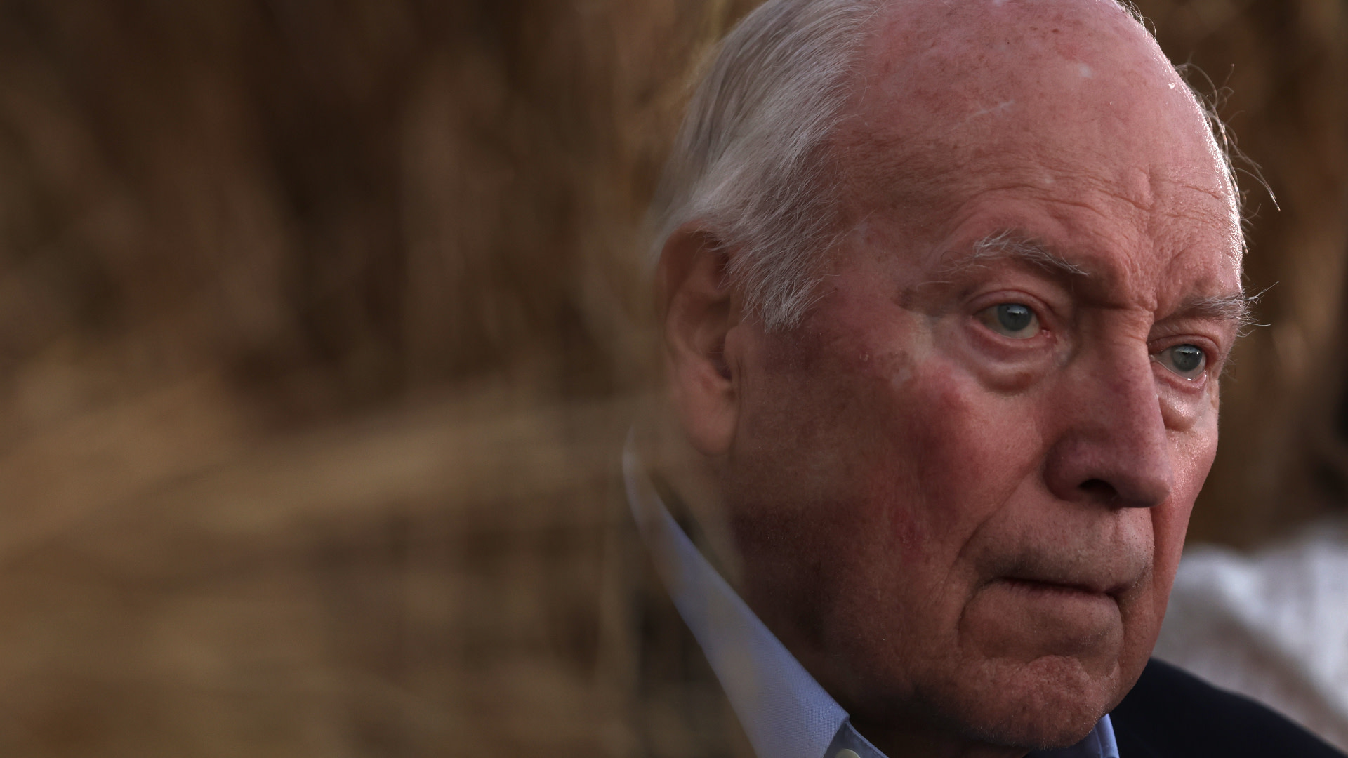 An elderly man with a serious expression, partially obscured by blurred foreground elements, in a natural setting.