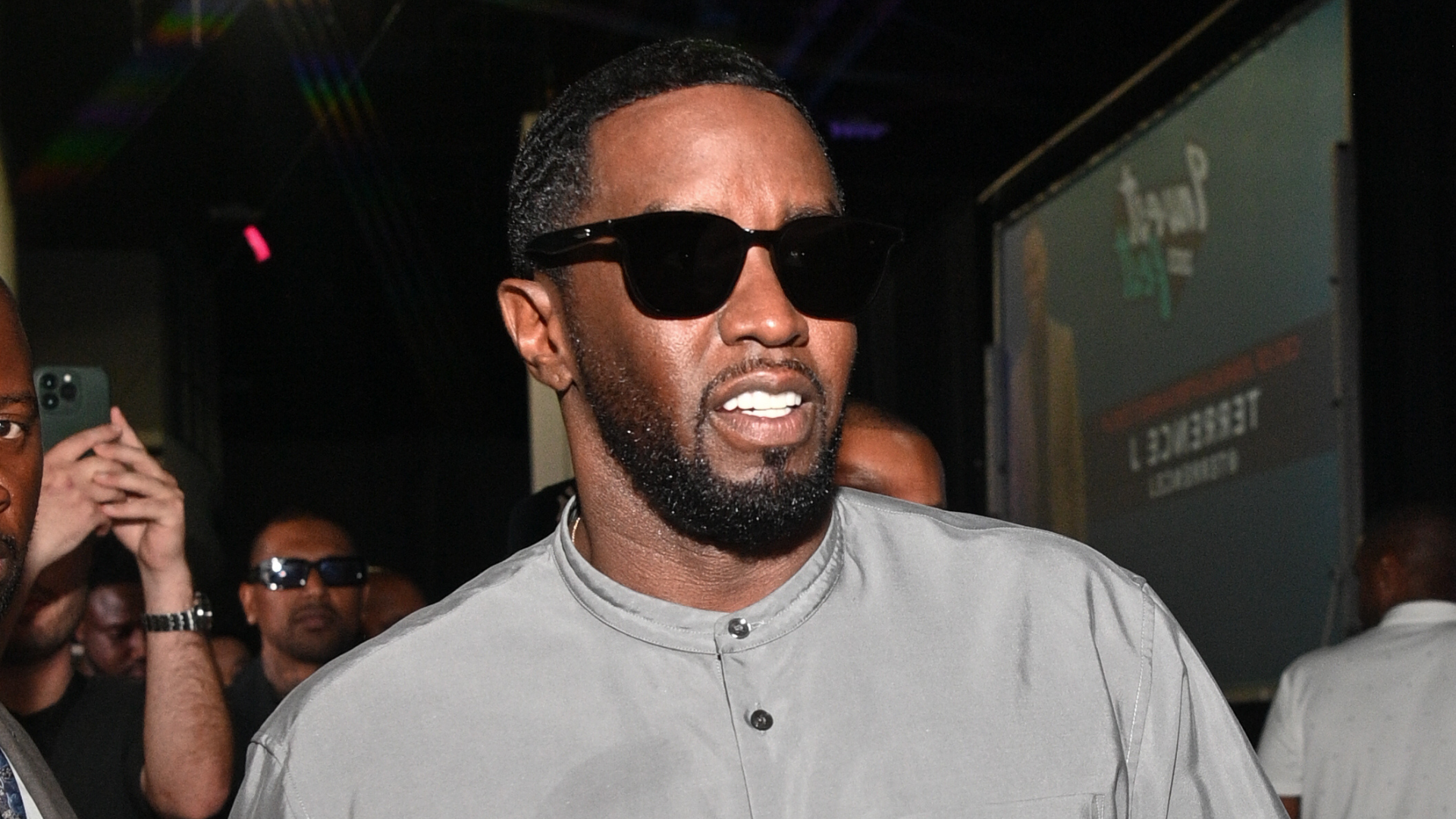 A man wearing sunglasses and a gray shirt, walking indoors with people in the background.
