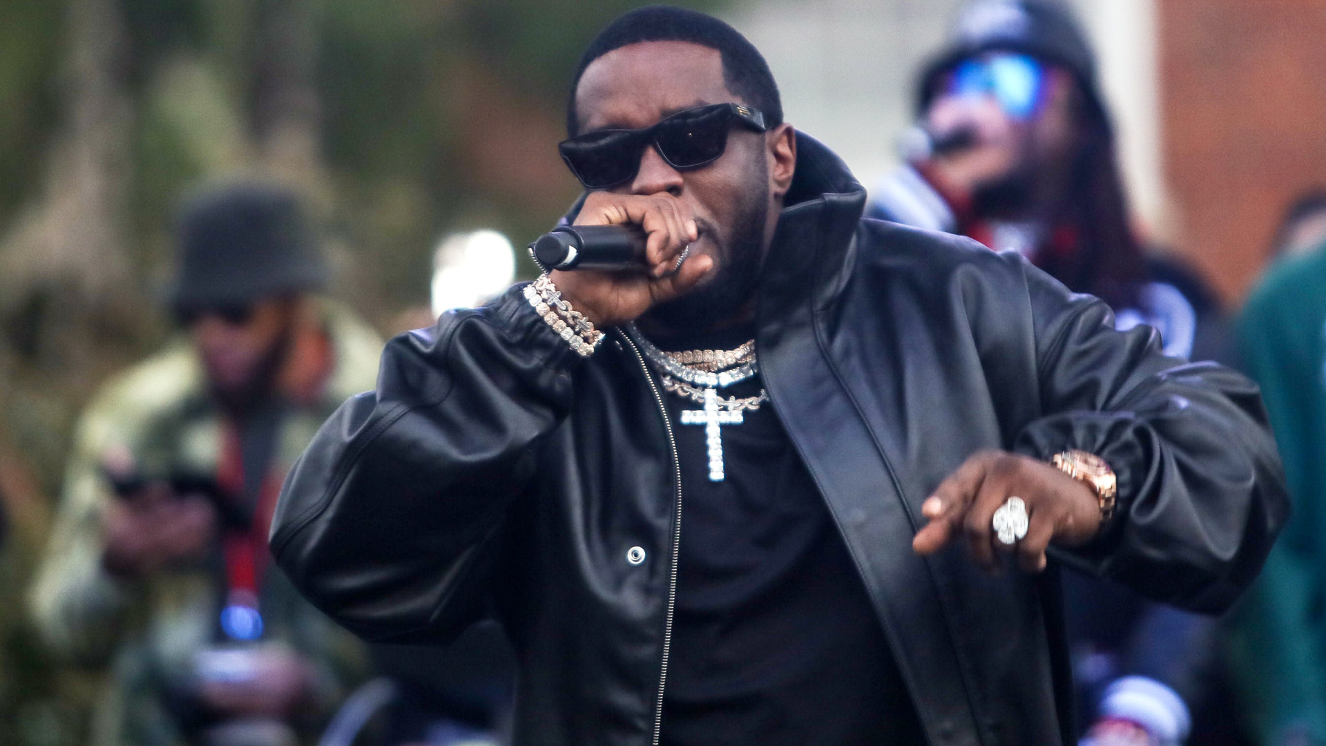 Diddy in a black leather jacket and sunglasses performs on stage, holding a microphone. He is wearing jewelry and has a confident expression.