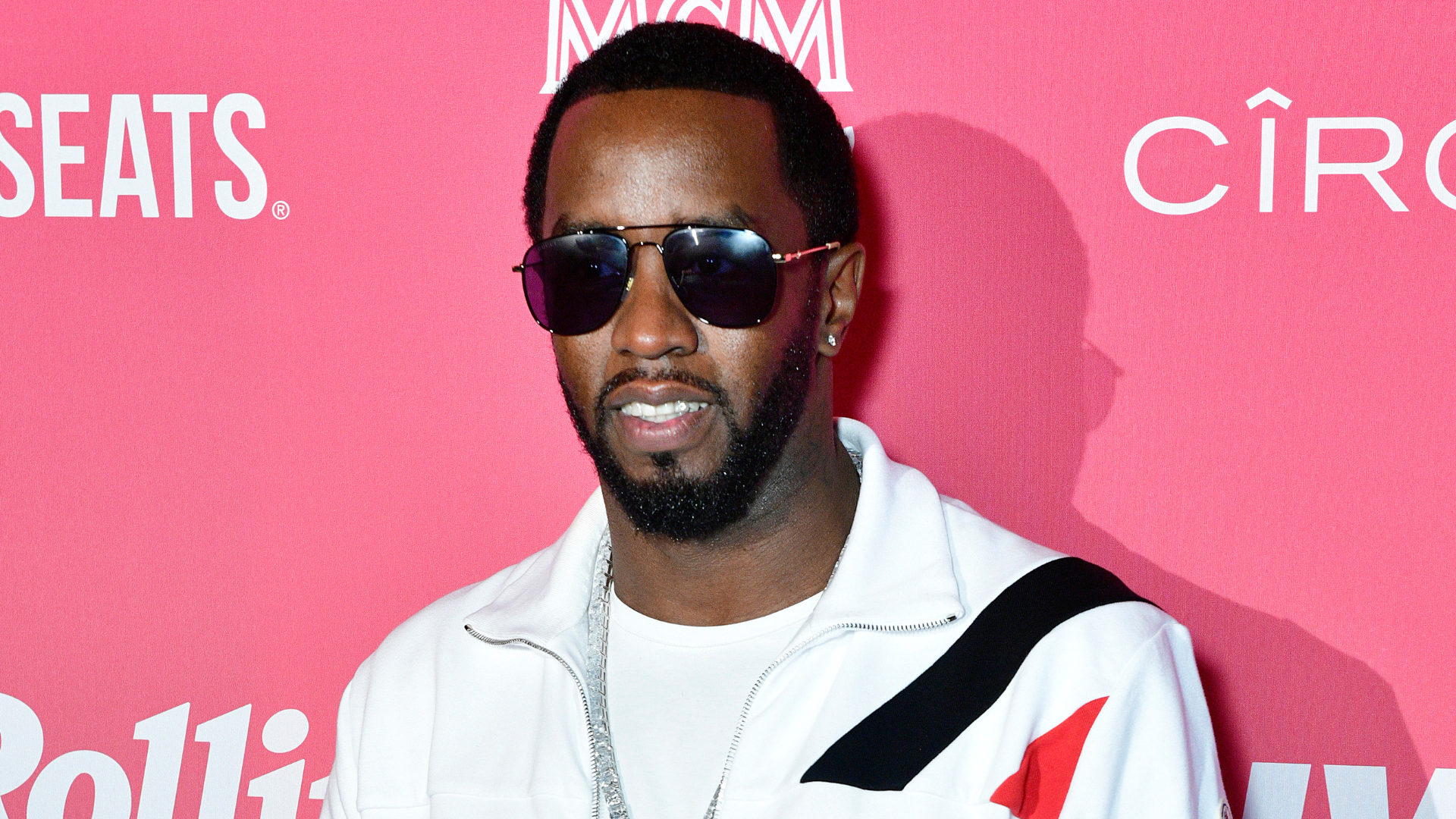 Sean "Diddy" Combs wearing sunglasses, a white jacket, and a chain, stands against a pink background with logos.