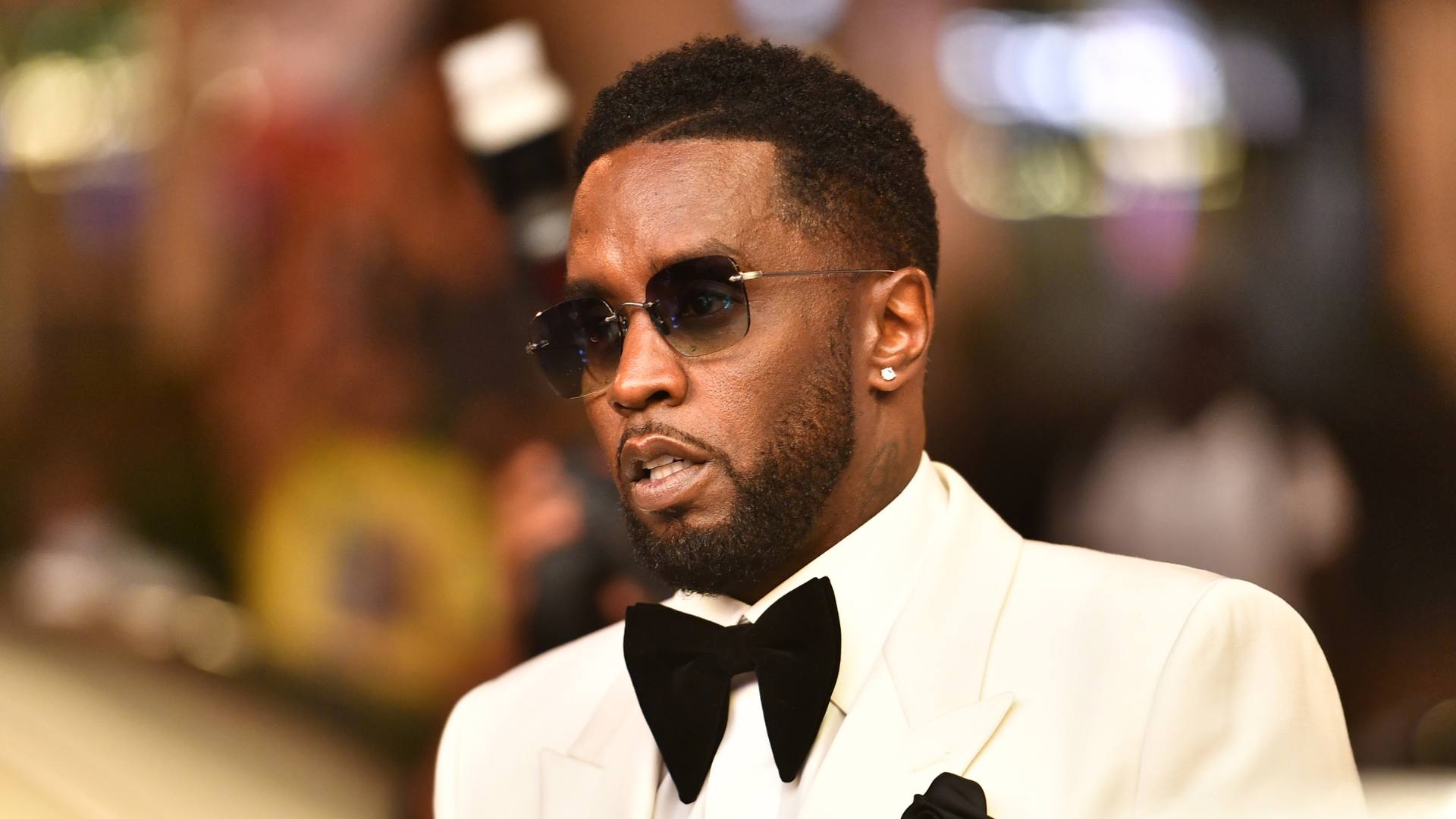 Sean "Diddy" Combs in a white tuxedo with a black bow tie, wearing sunglasses, at a formal event.