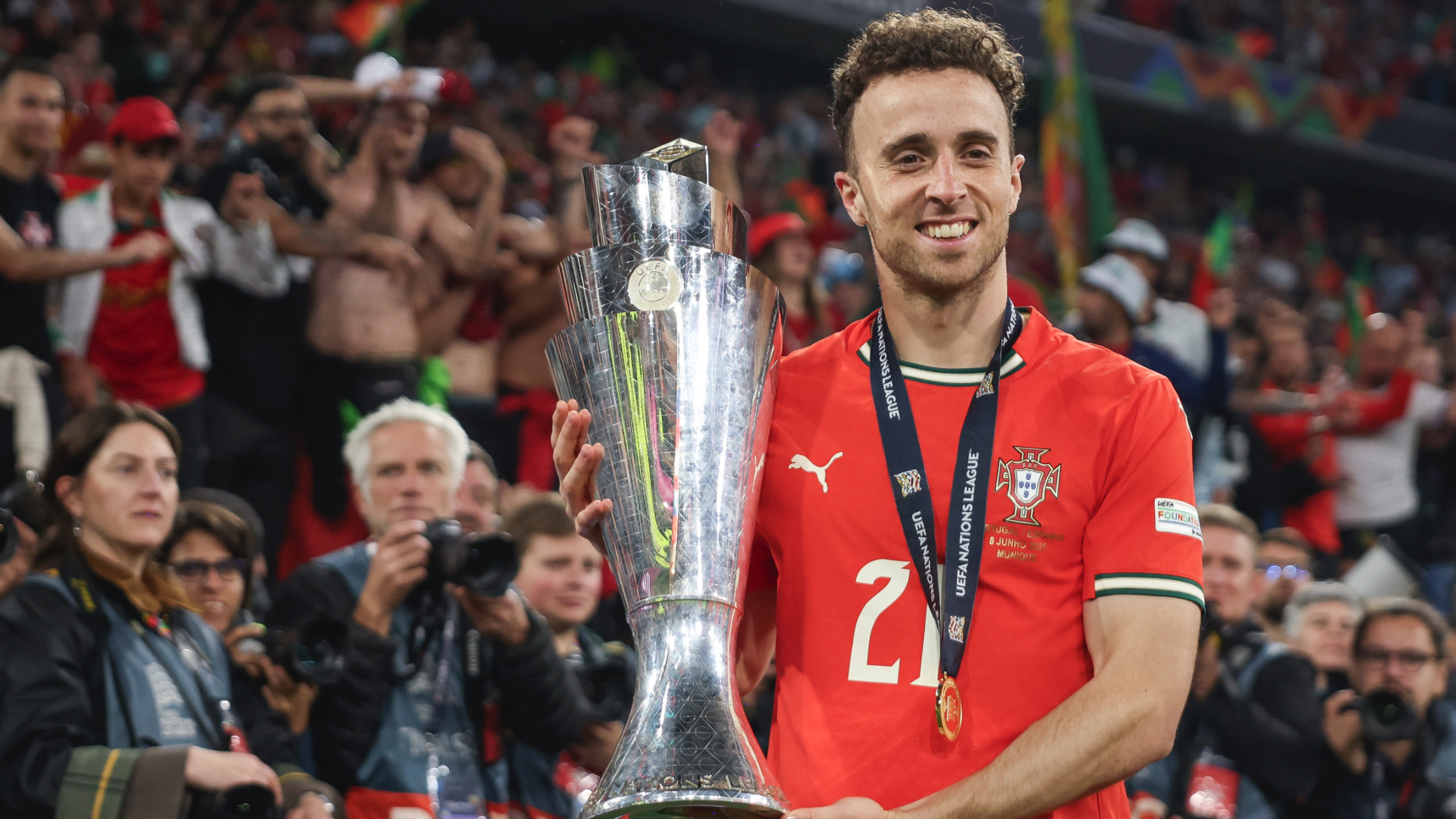 Diogo Jota in a red Portugal jersey holds a large trophy, smiling. The crowd is cheering in the background.
