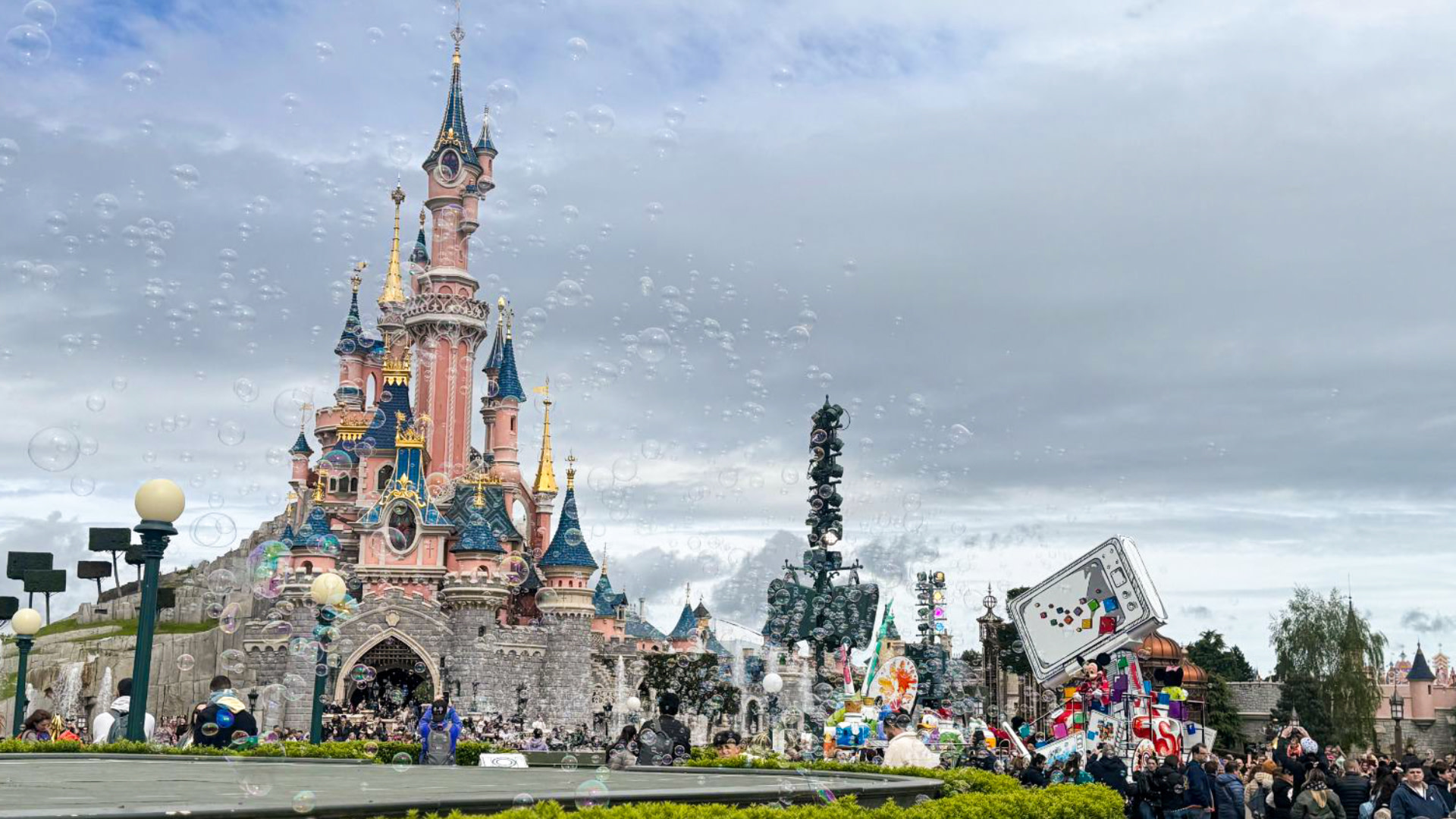 A colorful parade at Disneyland Paris with Sleeping Beauty Castle in the background, surrounded by bubbles and a crowd of visitors.