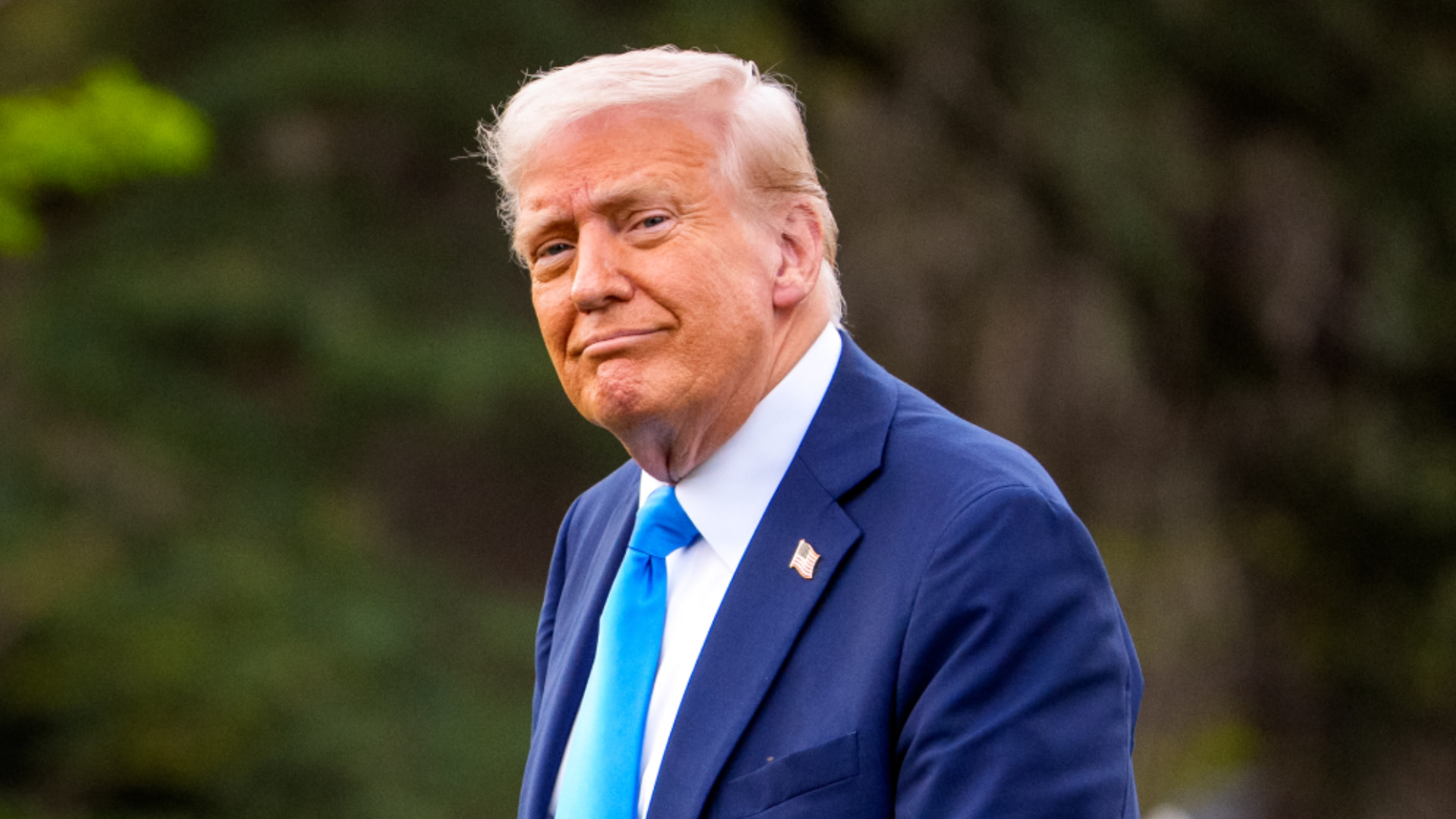 Donald Trump in a blue suit and blue tie, standing outdoors with a slight smile.