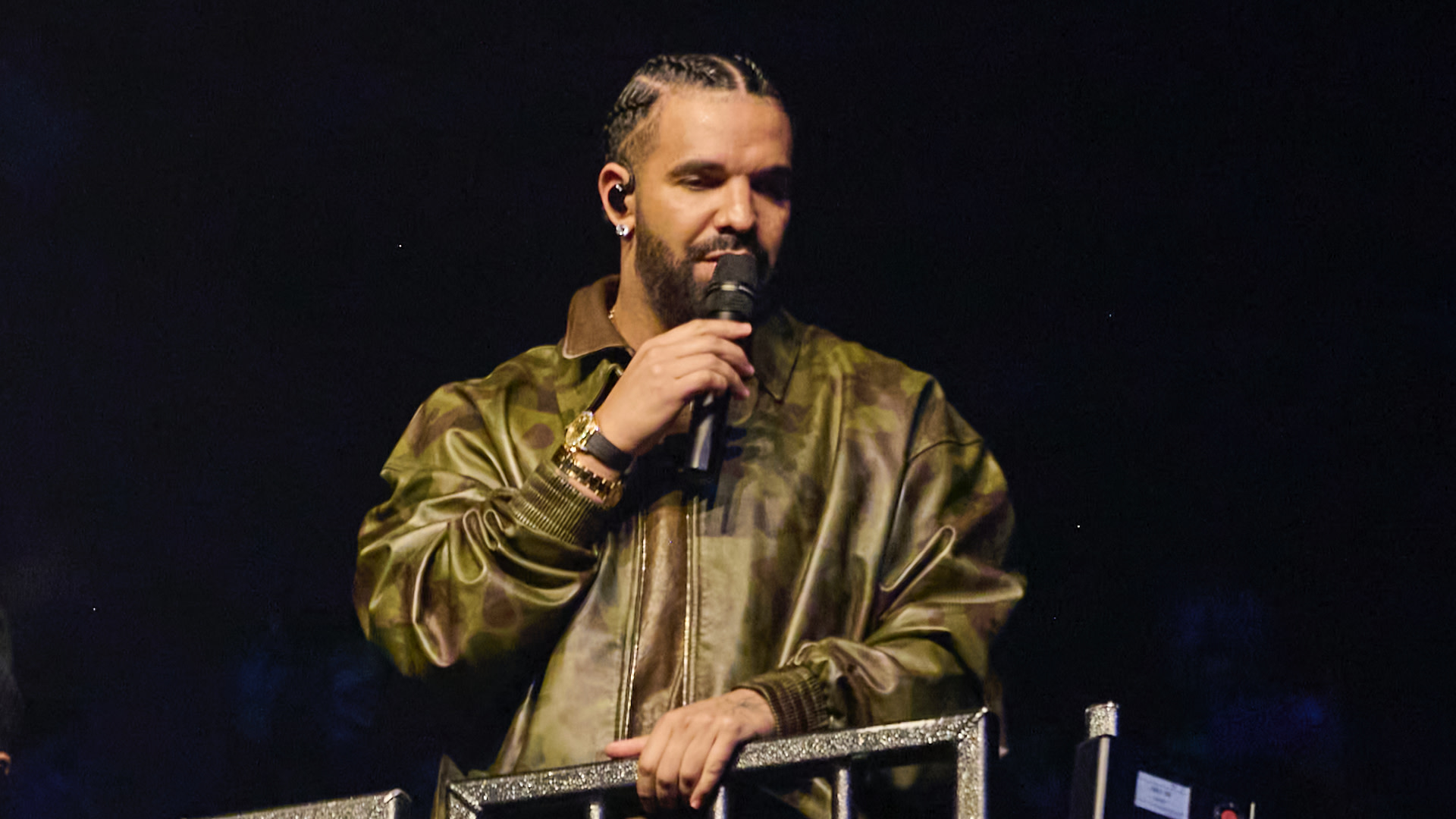 Drake performing on stage, wearing a camo jacket and holding a microphone.