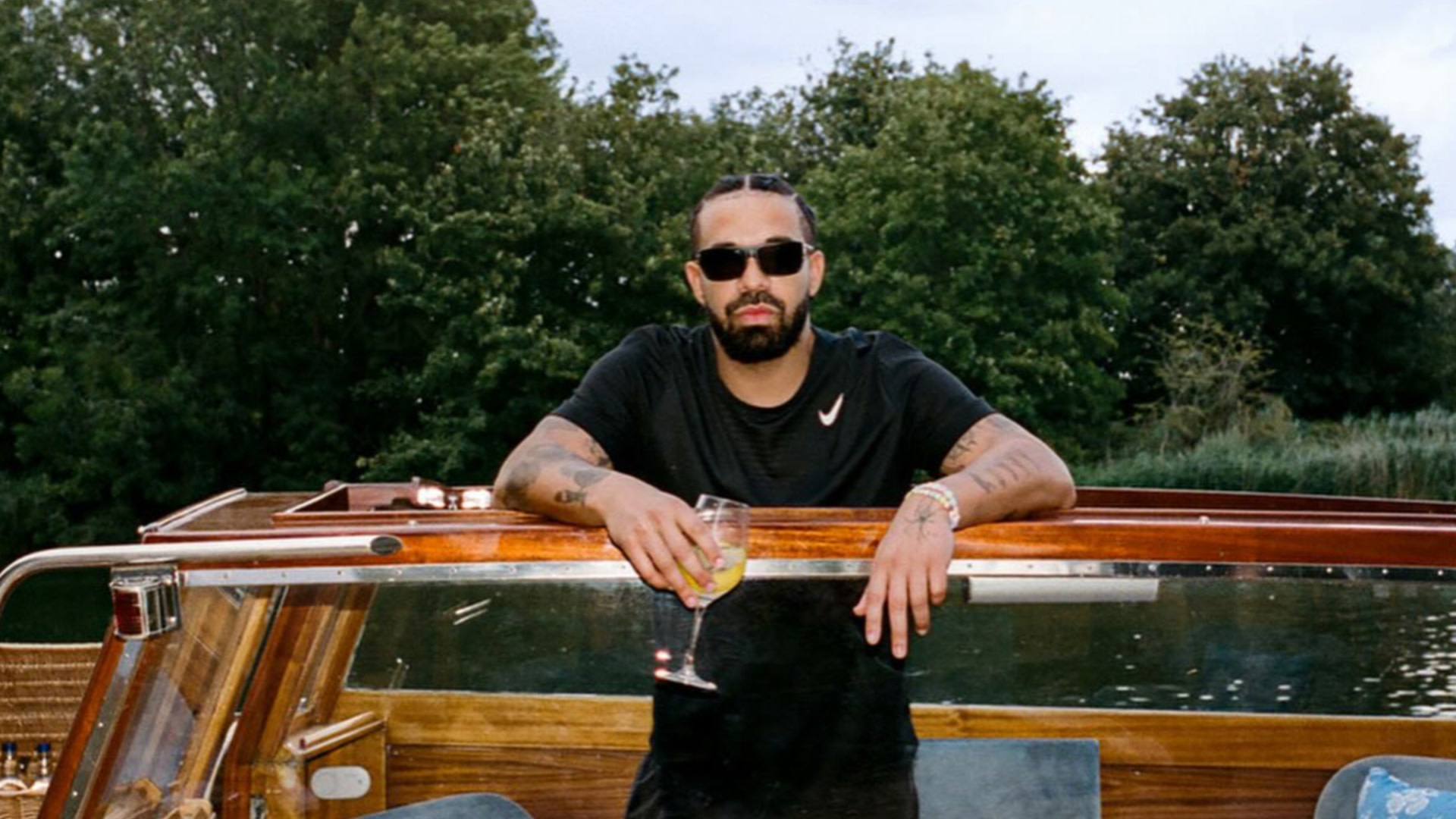Drake with sunglasses and braids holding a drink on a wooden boat, surrounded by trees.