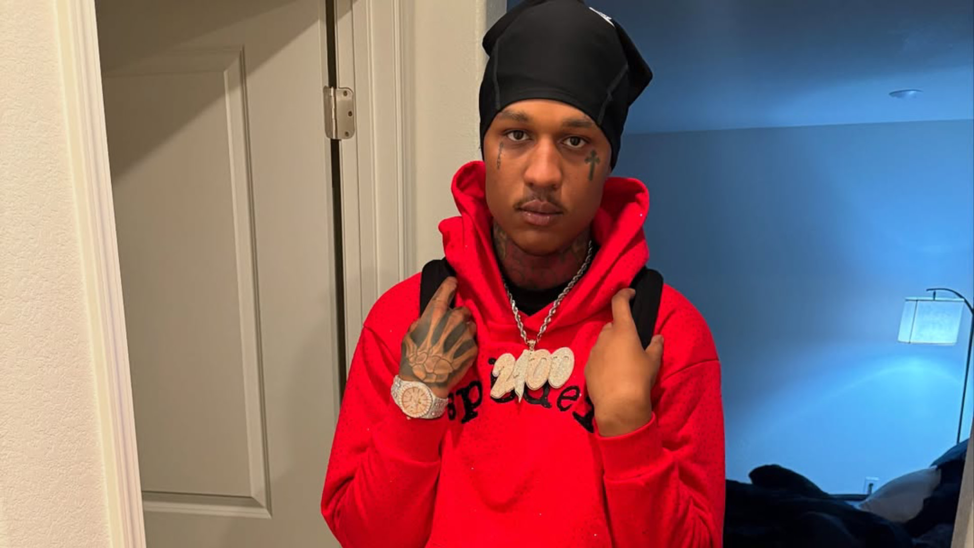 EBK Jaaybo in a red hoodie and black headscarf stands indoors, wearing a watch and necklace.