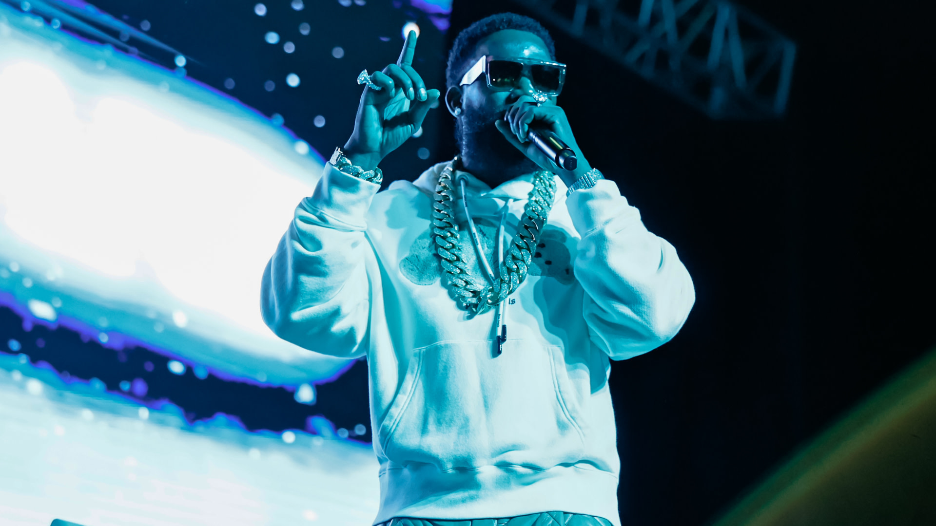 Gucci Mane on stage wearing sunglasses, a white hoodie, and heavy gold chains, holding a microphone with a vibrant background.
