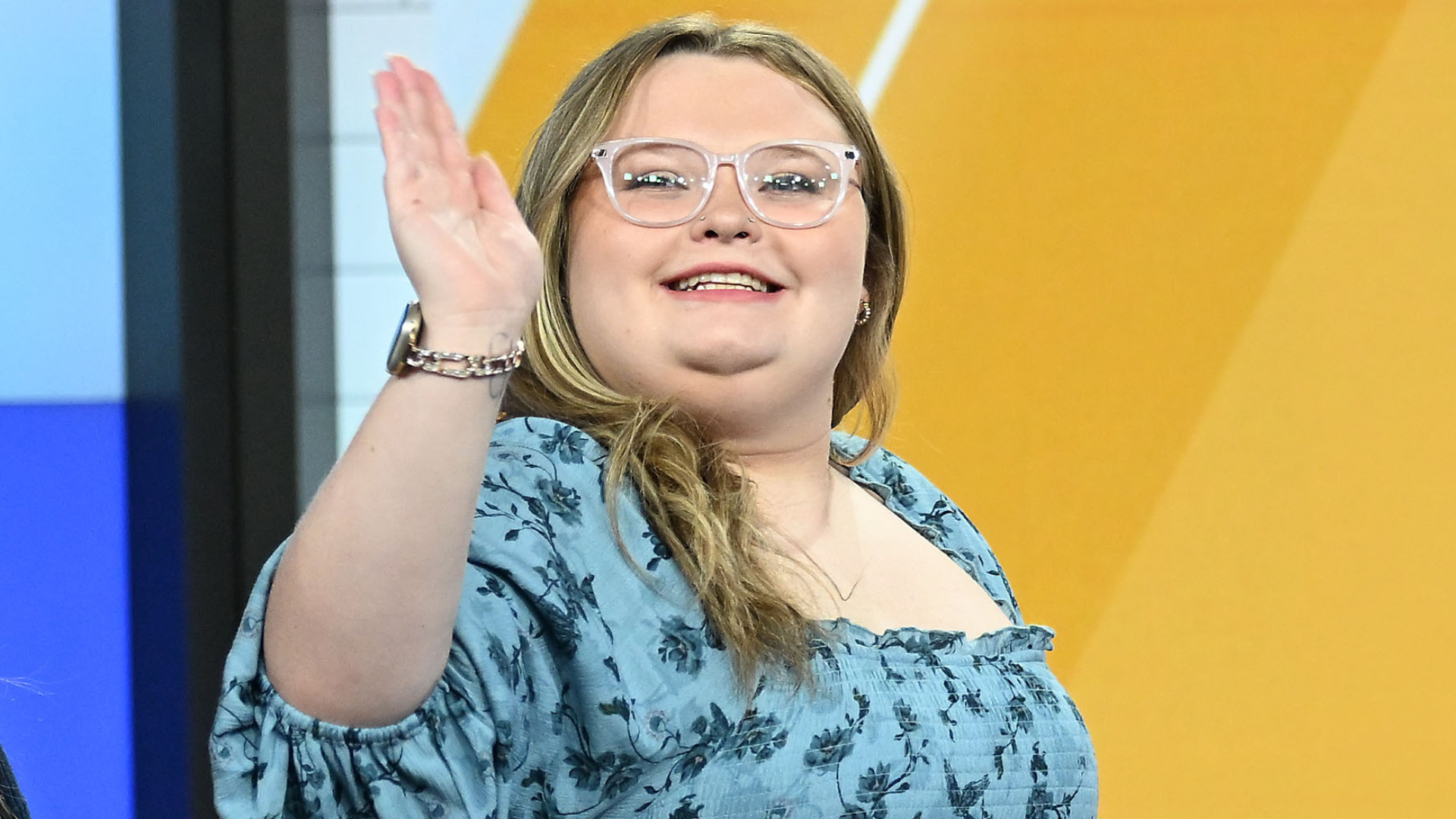 A person with glasses and a blue floral top waves, smiling against a yellow background.