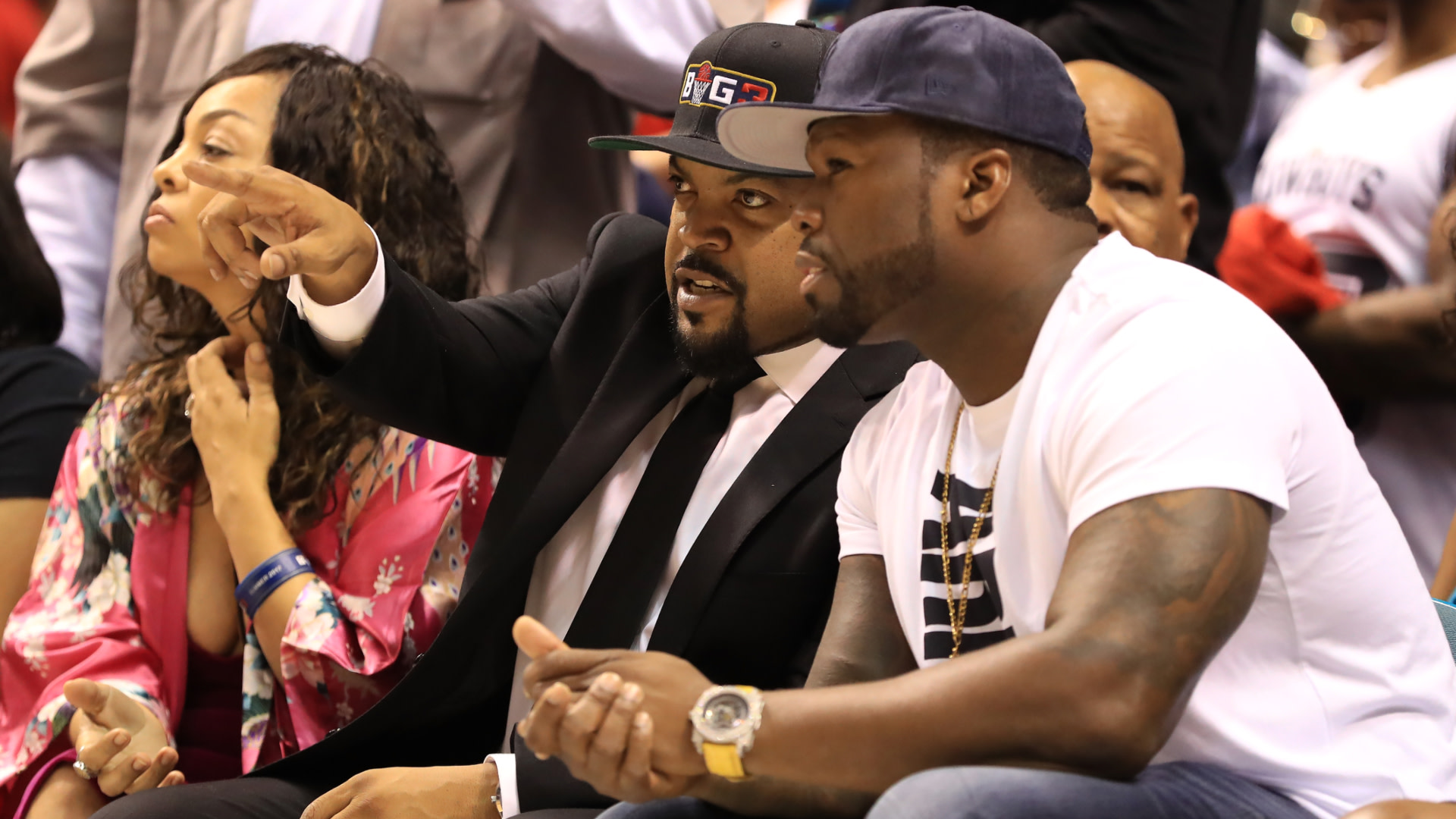 Ice Cube in a suit and cap points while sitting next to 50 Cent in a white t-shirt and cap at an event.