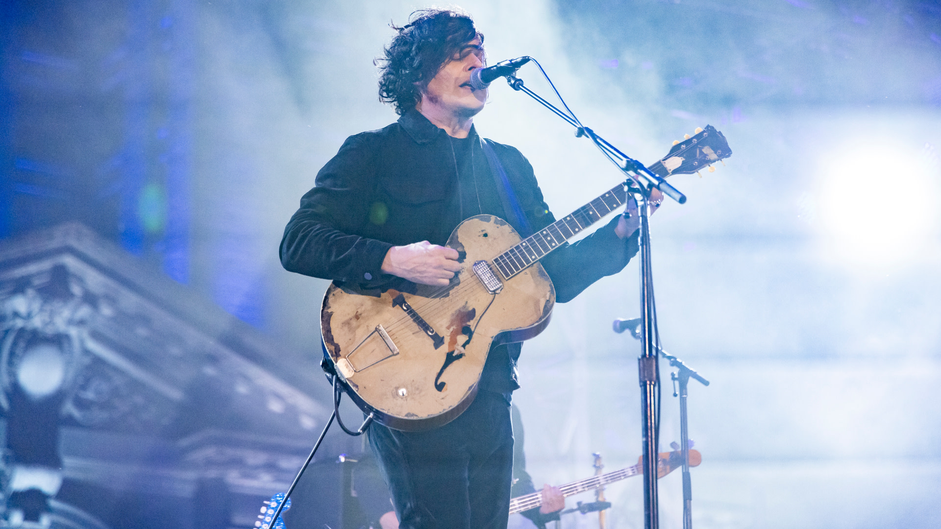 Jack White performing on stage with an electric guitar, wearing a dark outfit. The background is lit with blue and white stage lights.