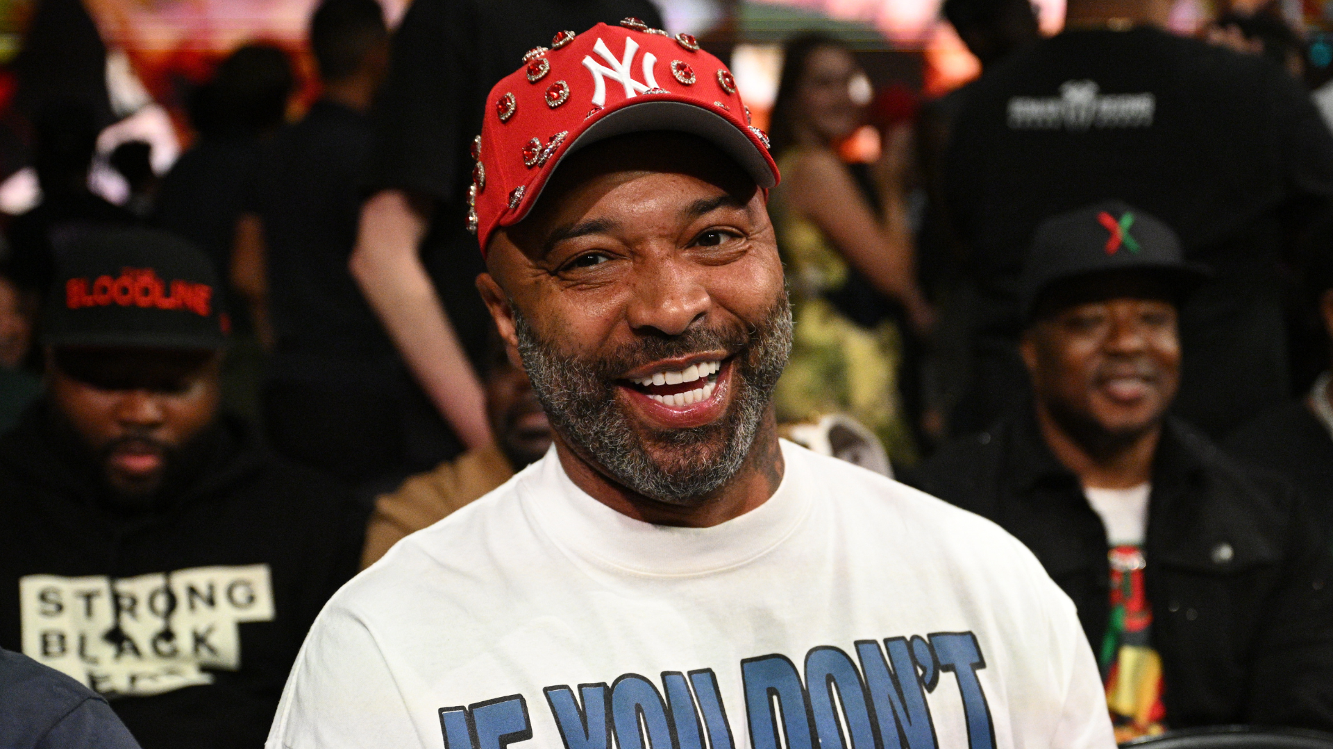 Man wearing a red cap with pins and a white shirt, smiling at an event with people in the background.