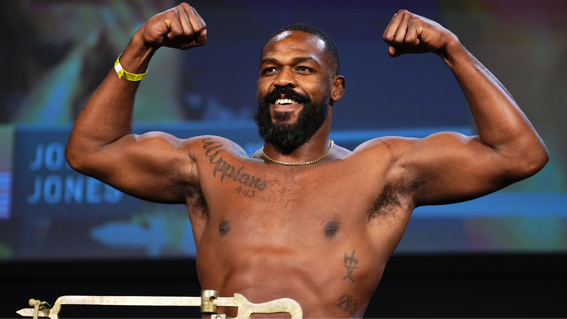 A muscular man with tattoos flexing his arms and smiling, standing on a weigh-in stage.