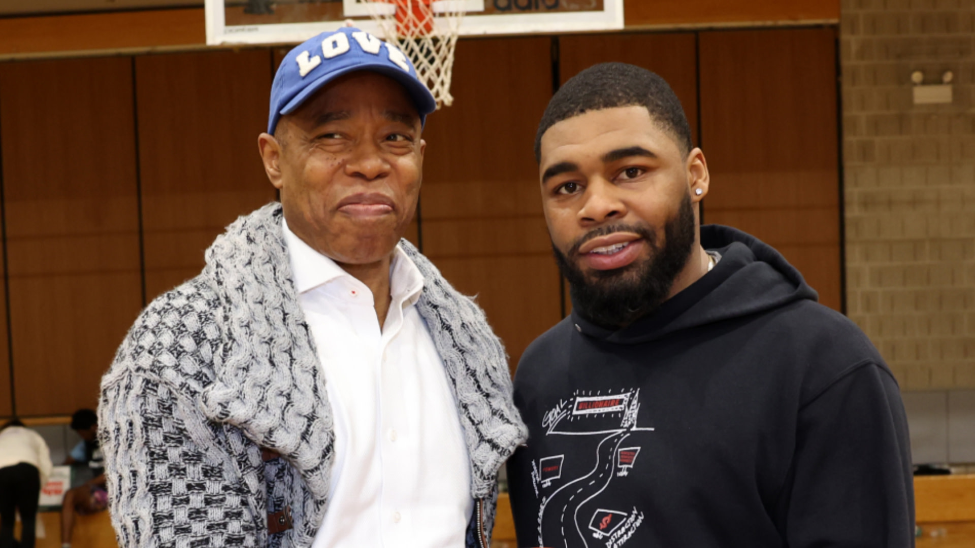 Mayor Eric Adams and son Jordan Coleman standing together indoors. Adams is wearing a blue cap and a patterned sweater, while Jordan is in a black hoodie.