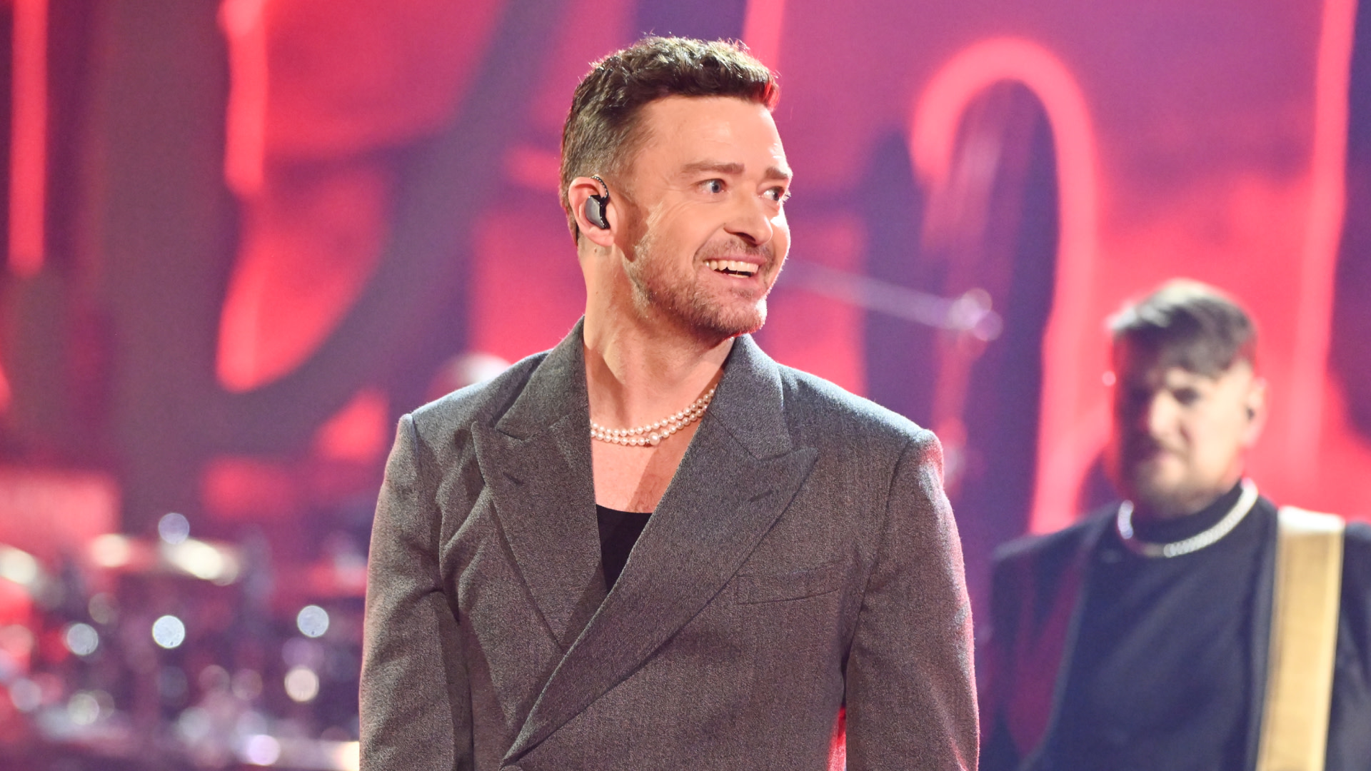 Justin Timberlake performing on stage, wearing a gray suit and pearl necklace, with a red background.