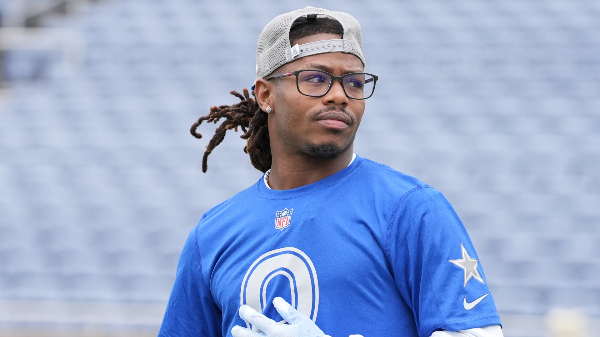 A man in a blue NFL jersey and cap stands on a football field, looking sideways.