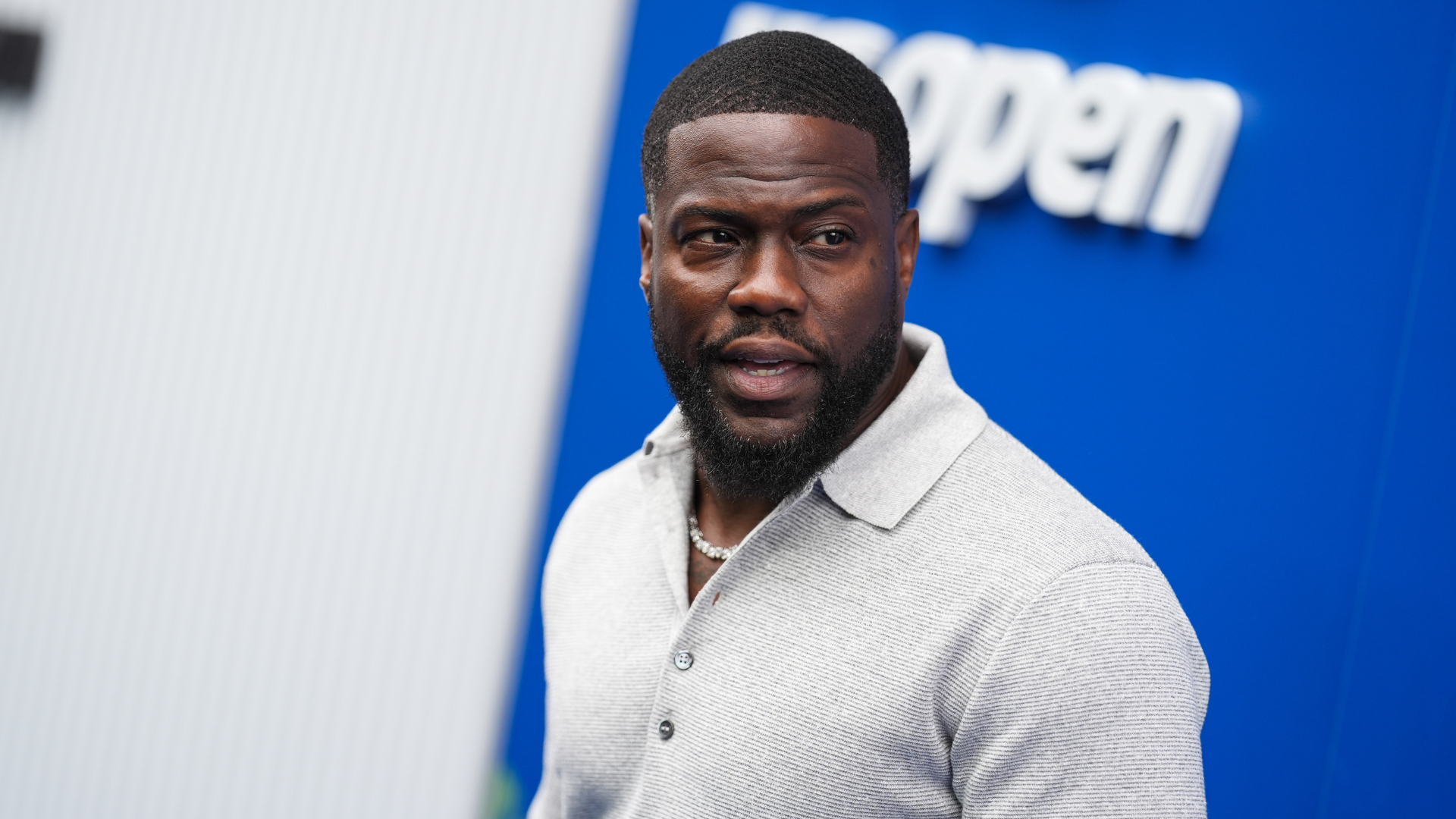 Kevin Hart in a light gray shirt, standing in front of a blue background with the word "open" partially visible.